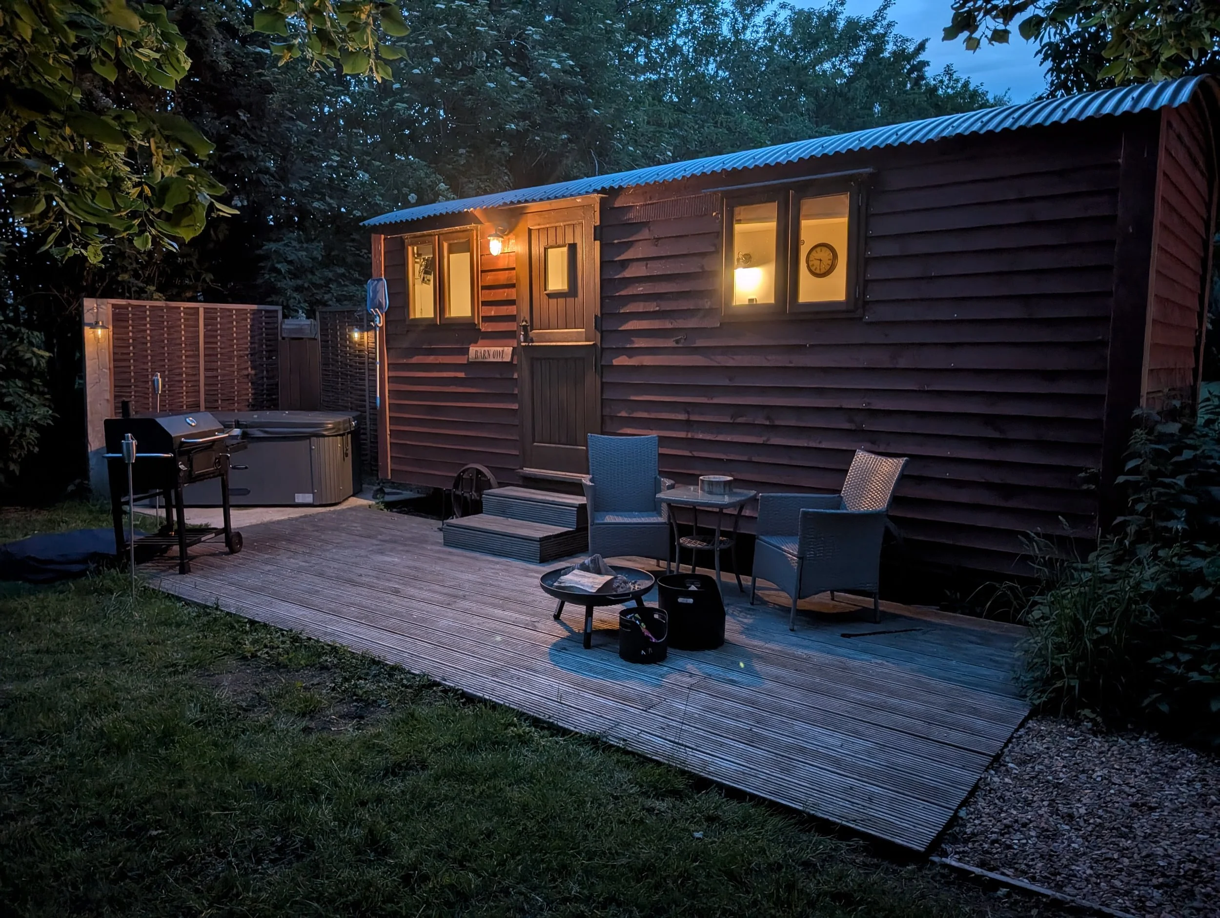 A small wooden cabin with lights on, seen at dusk or early evening, surrounded by trees. There are outdoor chairs, a table, and a BBQ grill on a wooden deck in front of the cabin.