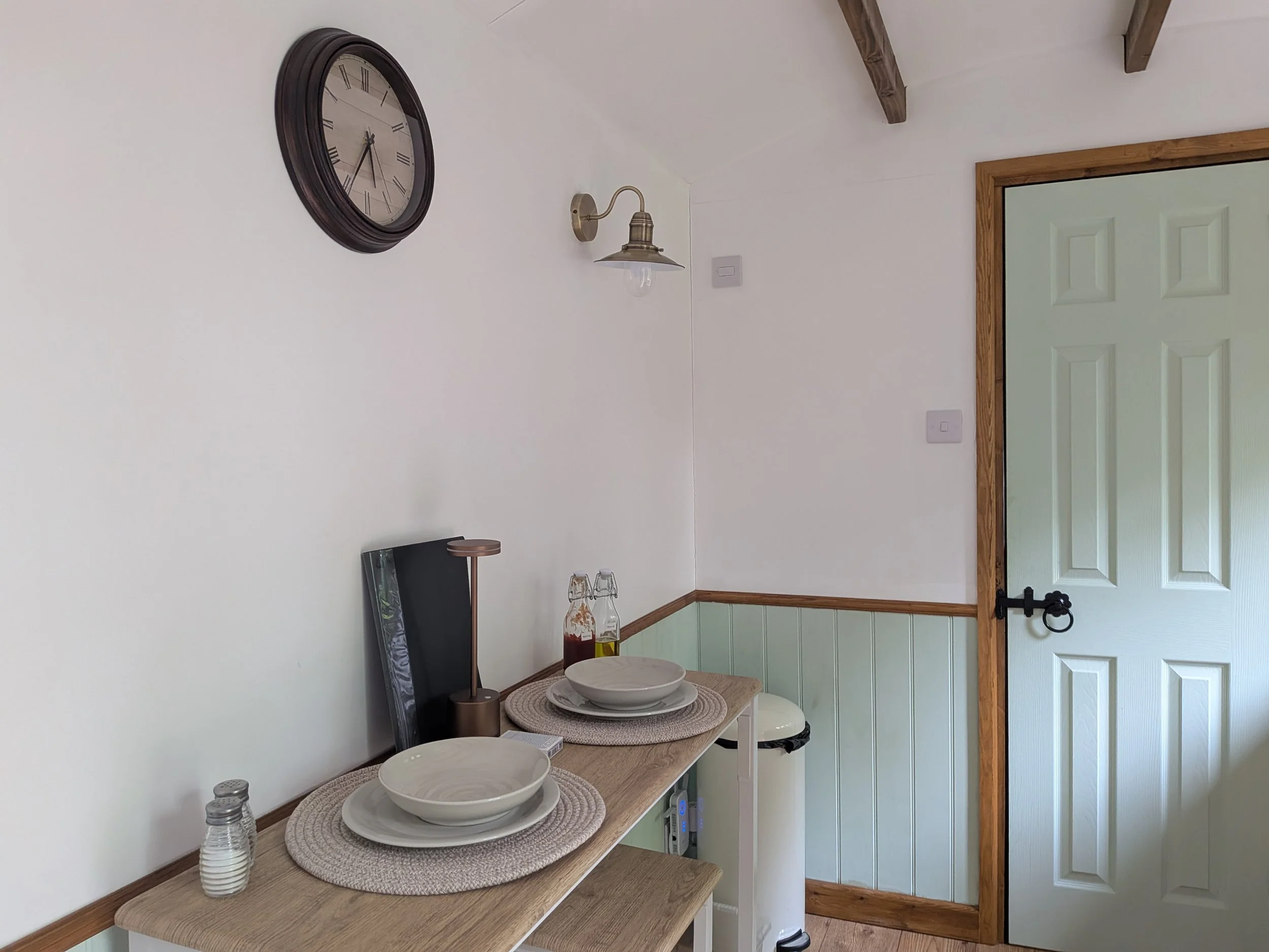 A dining area with a wooden table set with white bowls and placemats, two glass water bottles, and a small lamp on a side table. There is a large wall clock and a wall-mounted lamp, a door with a wooden frame, and light switches on the wall.