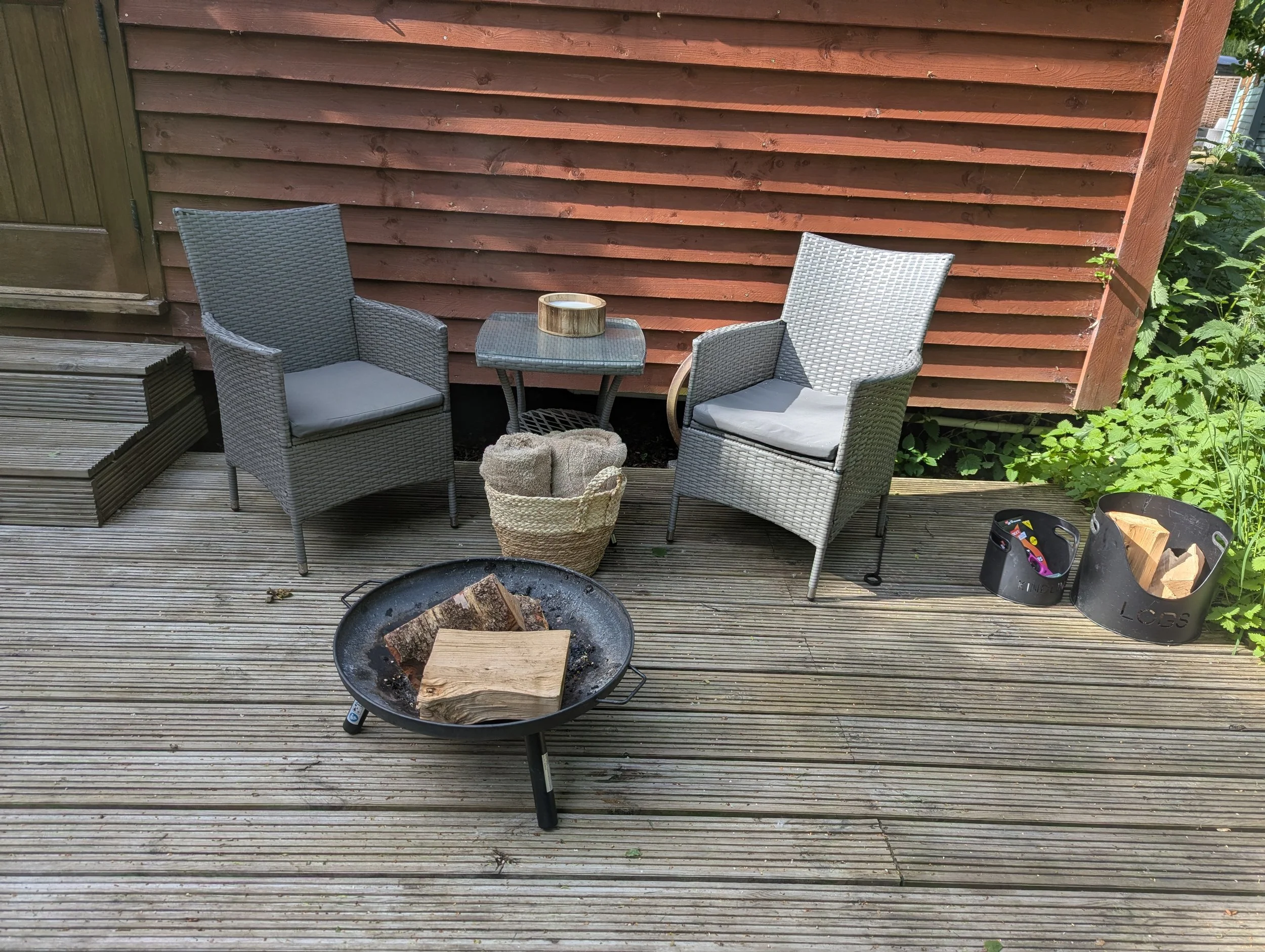 Backyard wooden deck with two gray wicker chairs, a small matching table, a basket, and a fire pit with logs. A red wooden fence and some green plants are in the background.