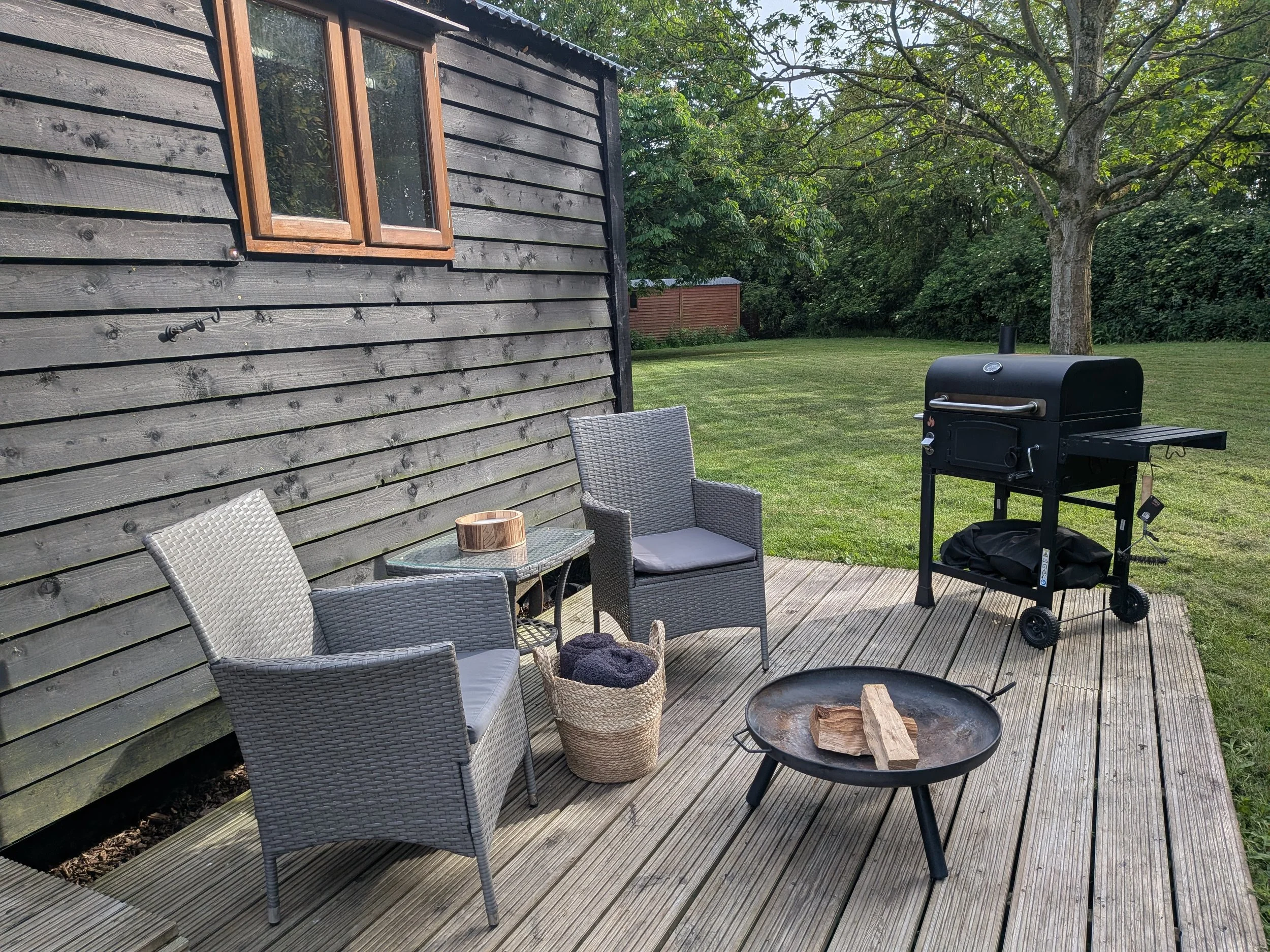 A backyard patio with a wooden deck, outdoor seating including two chairs and a table, a grill, and a fire pit with firewood, surrounded by green grass, trees, and shepherds hut.