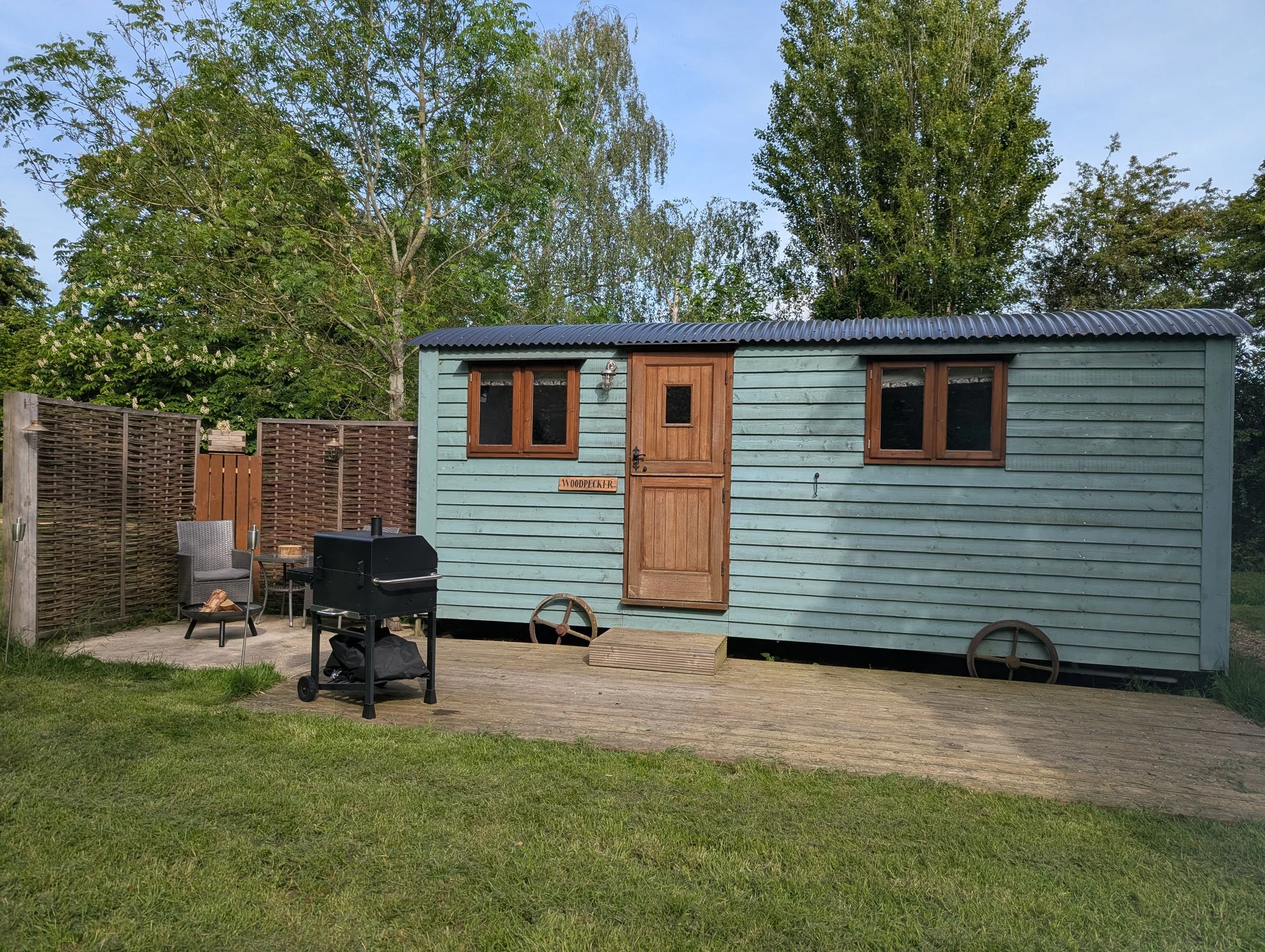 A green shepherd hut, two windows, and a door labeled 'Woodpecker,' situated in a backyard with a woven fence, outdoor furniture, a barbecue grill, and surrounded by trees and grass under a clear sky.
