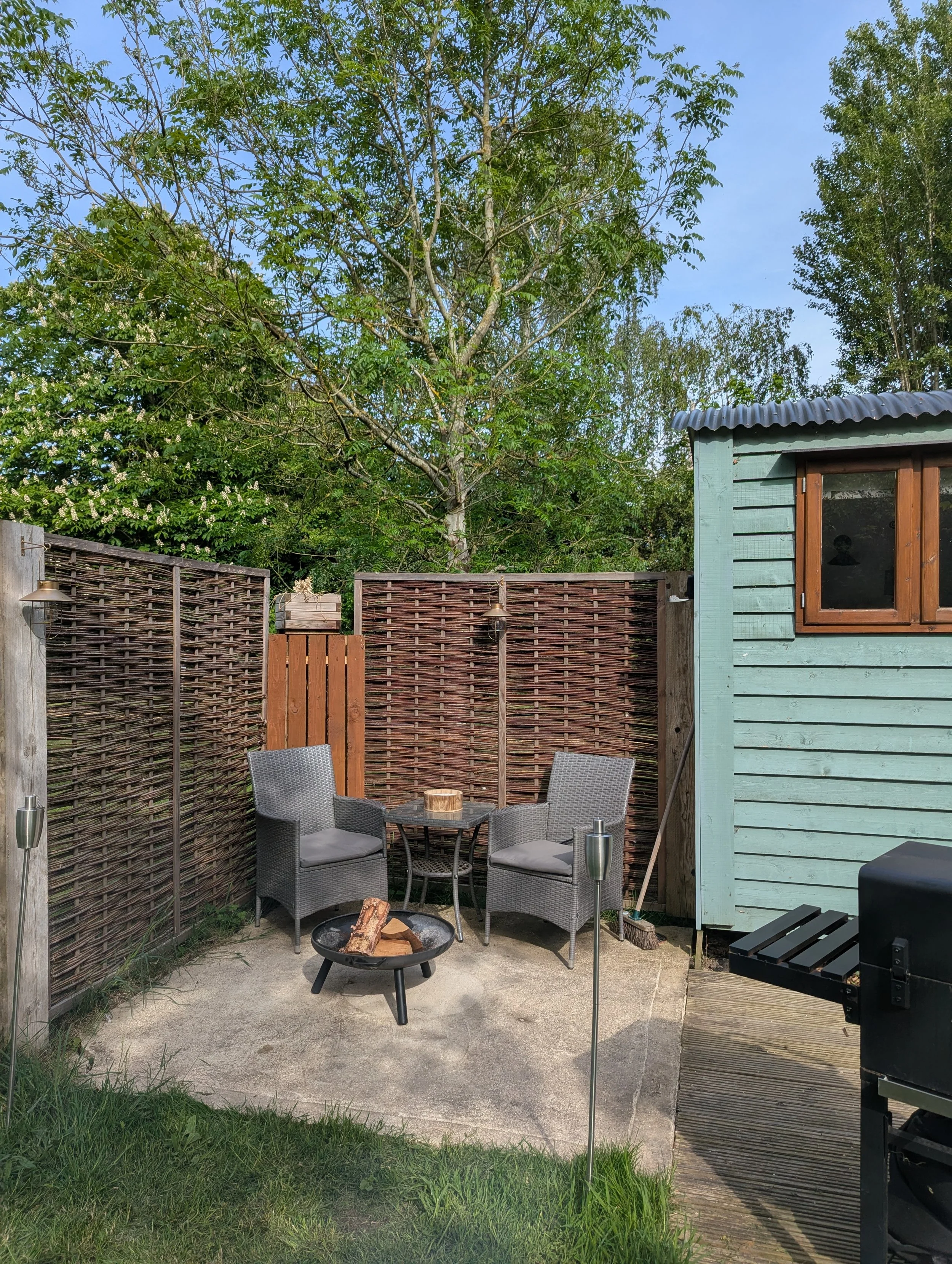A backyard patio with wicker chairs around a fire pit, a wood fence, a small green shed, and a large leafy tree in the background.