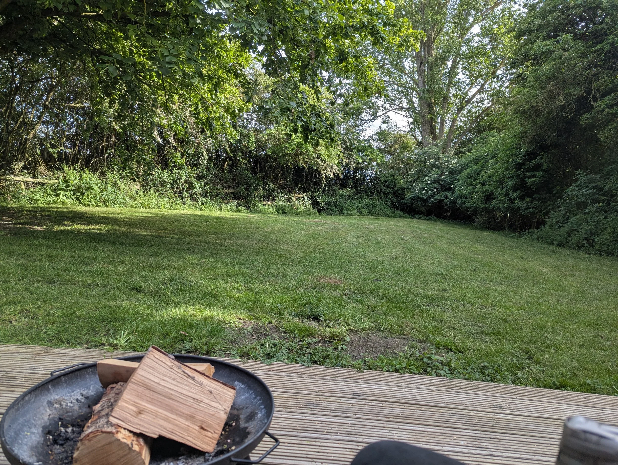 Firepit with several pieces of cut wood on a wooden deck, with a grassy yard and dense green trees in the background.
