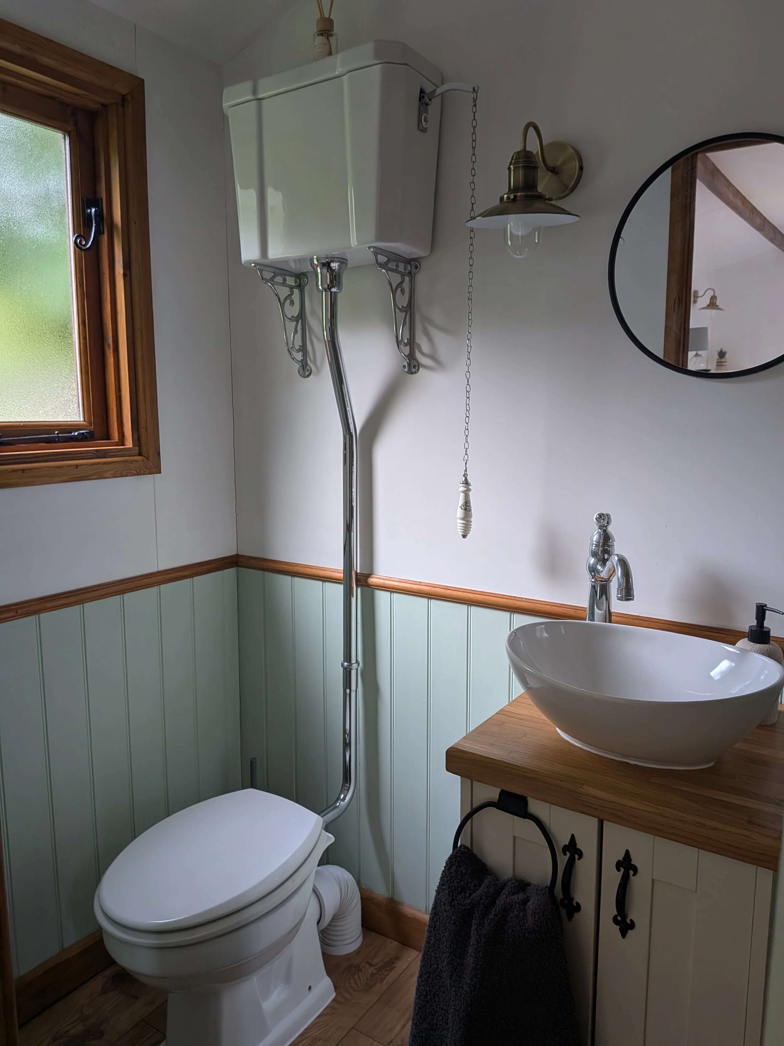A ensuite bathroom with a white toilet, a round vessel sink on a wooden countertop, a circular mirror, a window with wooden trim, and a brass wall-mounted light fixture. There is a black towel hanging on a decorative black iron hook.