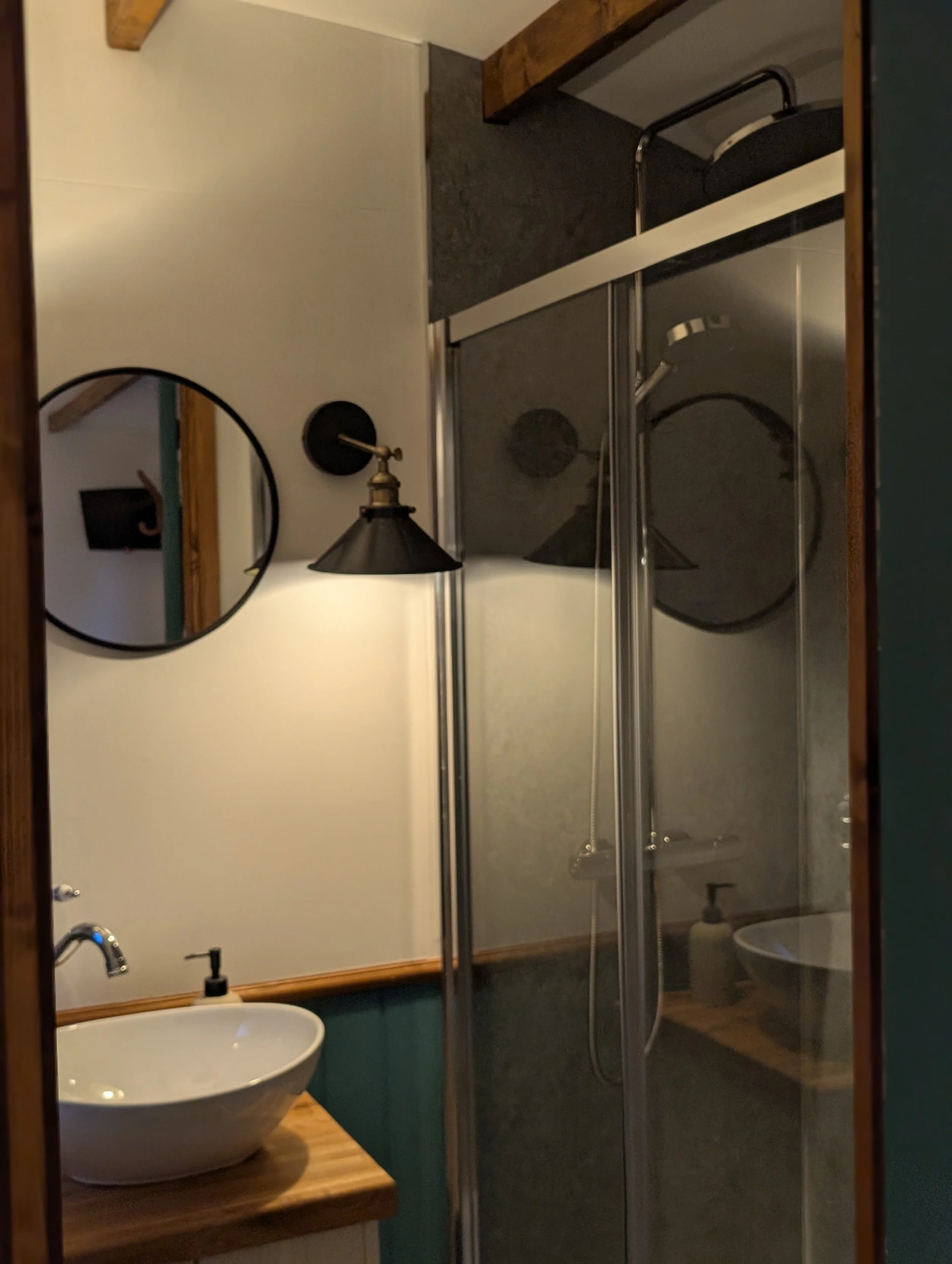 ensuite Bathroom with a round mirror, a wall-mounted light fixture, a wooden countertop with a vessel sink, and a walk-in shower with glass doors.