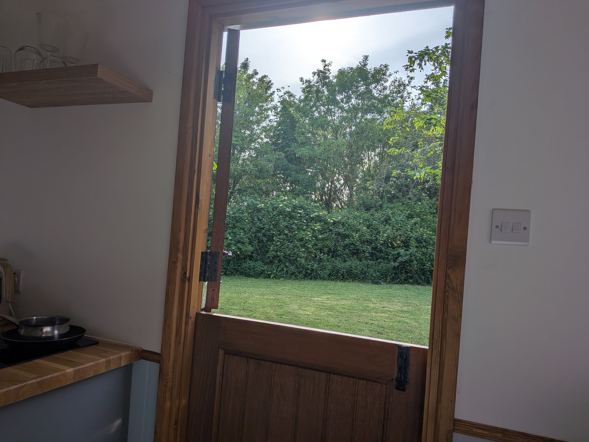 Open window with a view of green trees and grass outside, inside a shepherds hut with a wooden countertop and shelf with glasses.