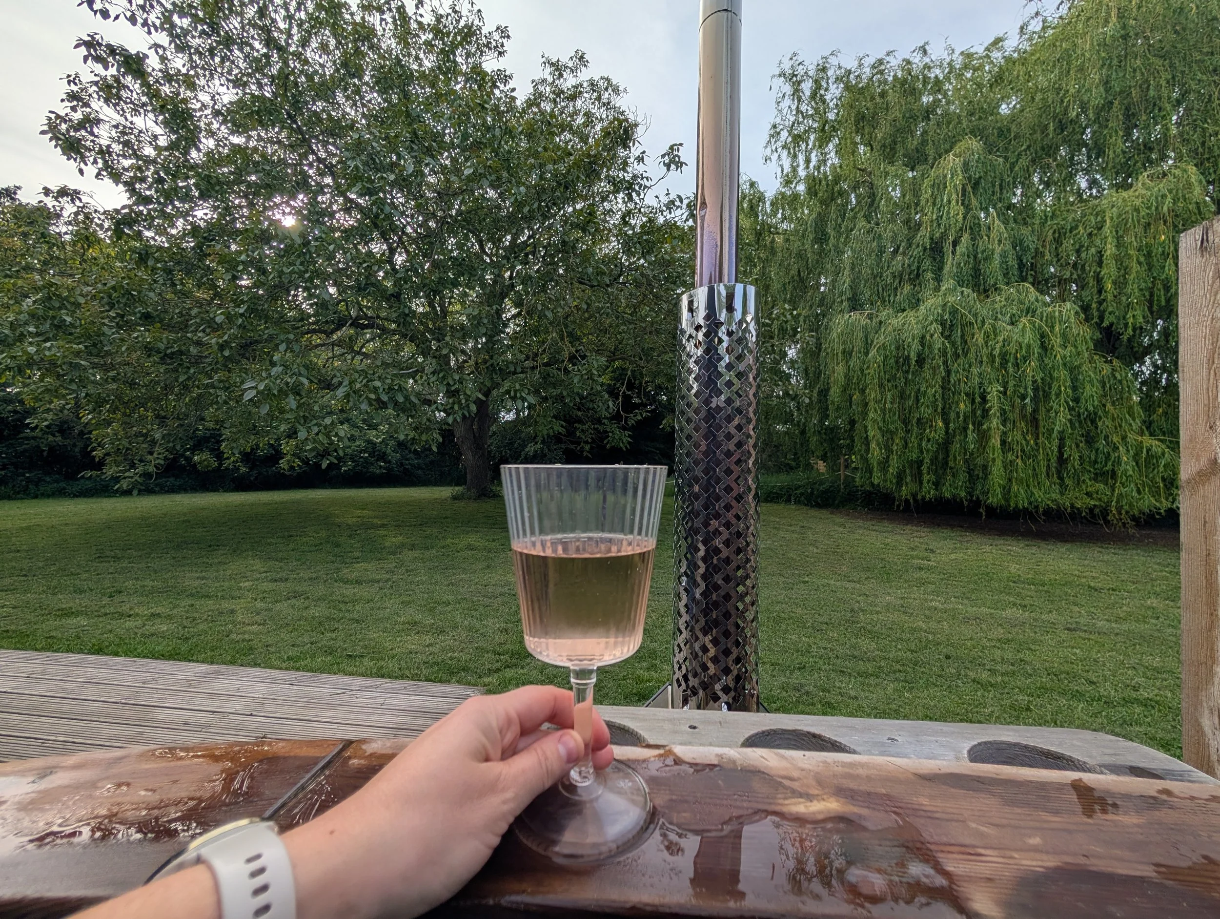 A person holding a glass of pink wine on an outdoor wooden hot tub, with a background of trees and a cloudy sky.