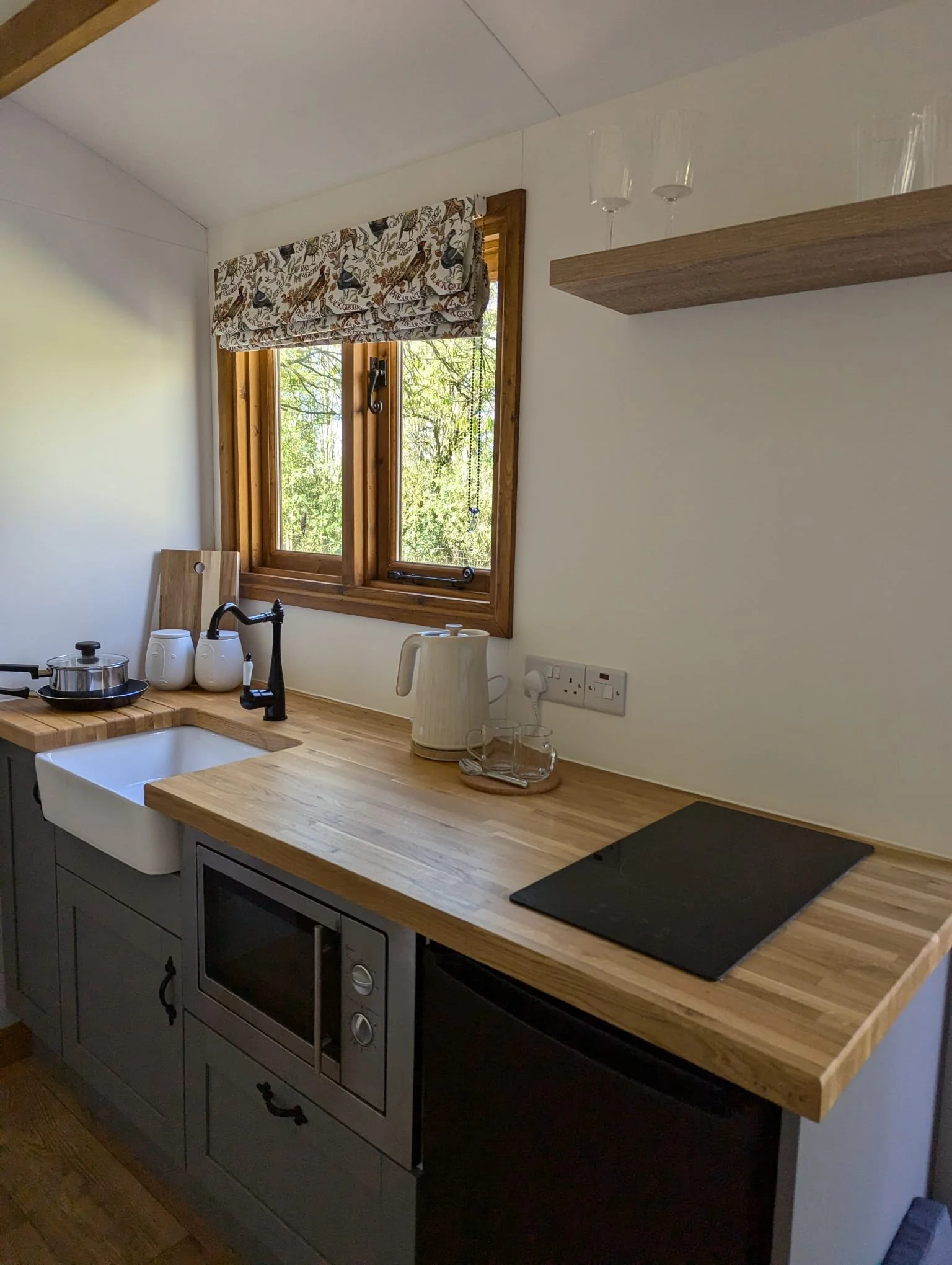 Kitchen with wooden countertops, a window with a patterned valance, and various kitchen items including a kettle, jars, a pot, and a black stovetop.