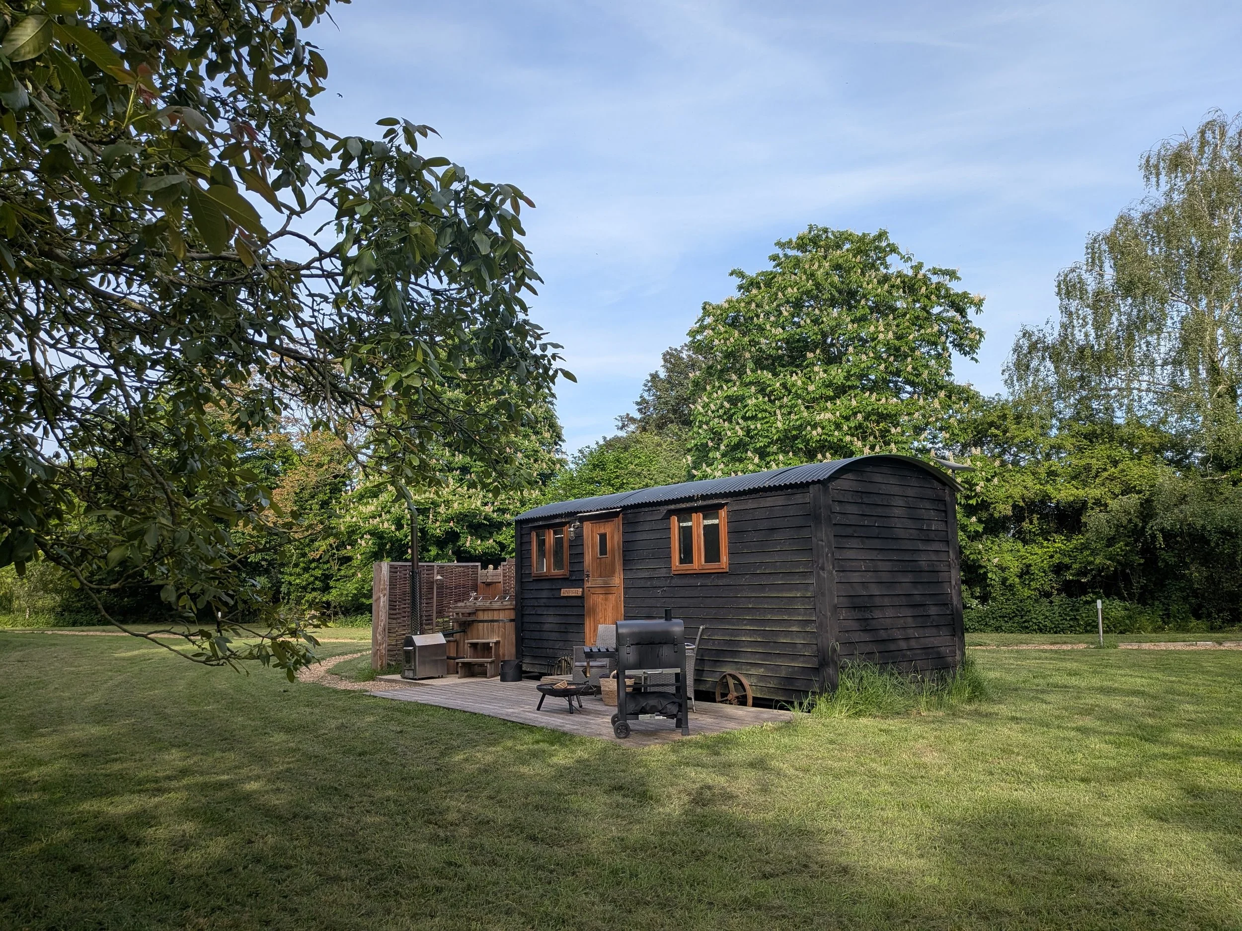 A black shepherds huts, located on a well-maintained lawn. In front, there are outdoor chairs and a barbecue grill. Tall green trees surround the area, and the sky is mostly clear with some thin clouds.