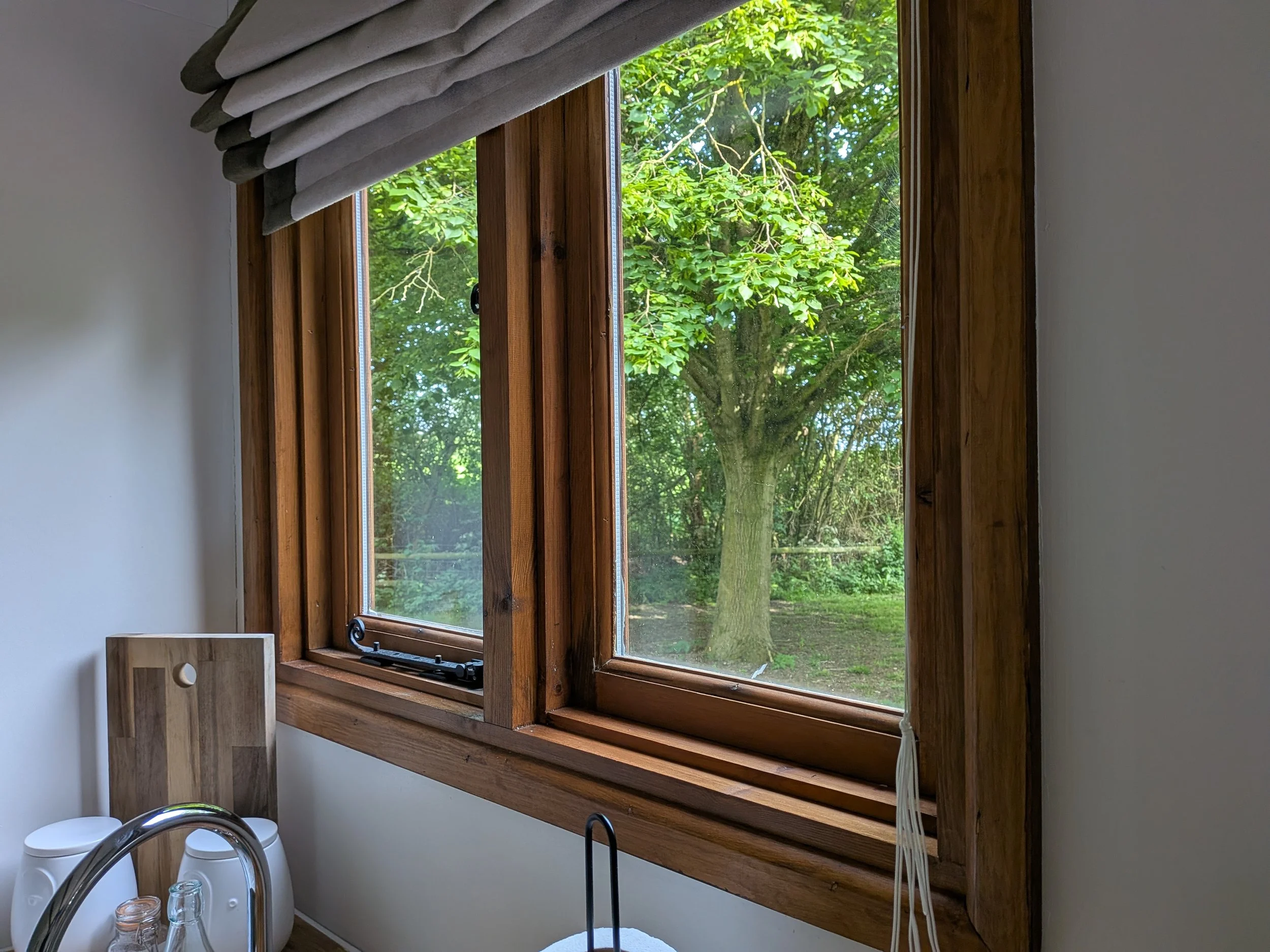 A woodworking window with wooden trim overlooking a lush green outdoor scene with trees, inside a room with part of a kitchen faucet and cups visible.