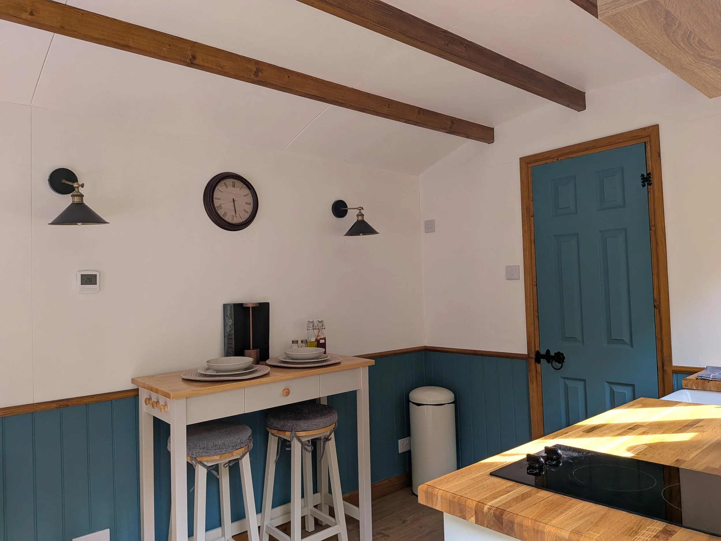 A cozy kitchen corner with blue paneled walls and a wooden countertop stove. A white table with two matching stools is placed against the wall, topped with ceramic dishes and placemats. The wall features a round wall clock and two black wall sconces 