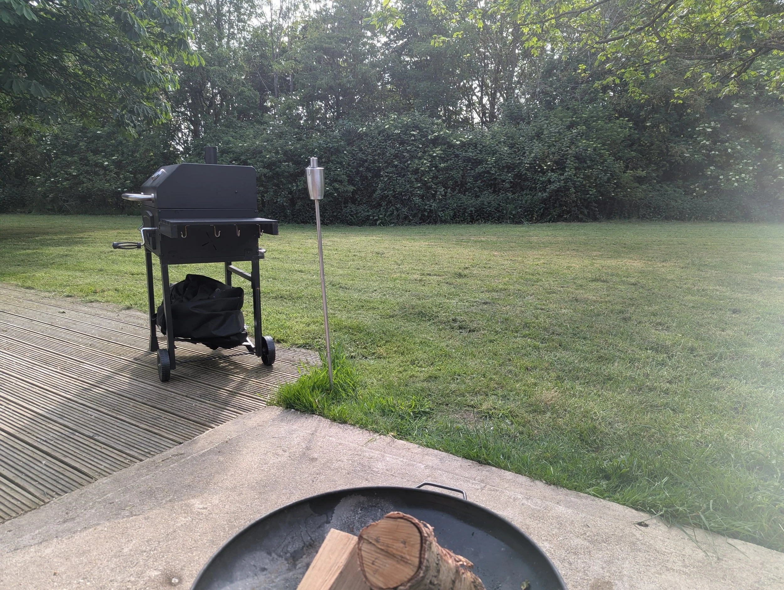 View from a person's perspective sitting outdoors near a fire pit with logs, looking at a grassy yard, bushes, and trees in the background, with a barbecue grill nearby.