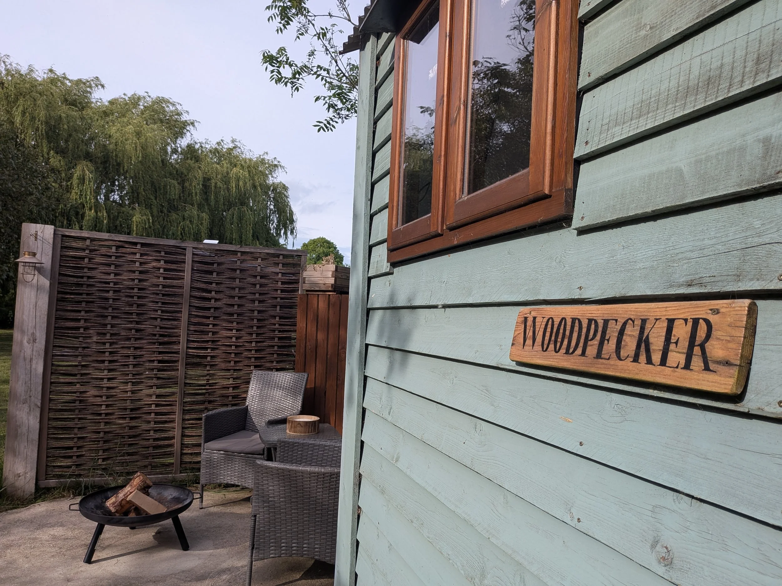 Outdoor patio with a light green wooden hut wall, a wooden window frame, patio furniture including two chairs and a table, a fire pit with logs, and a wooden fence in the background, surrounded by trees.