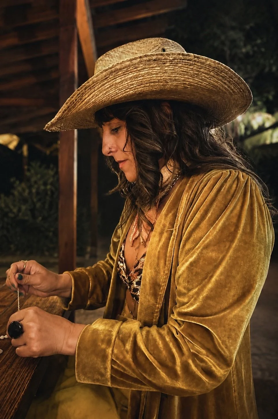 A woman wearing a large straw hat and an ochre velvet jacket, holding a jewelry chain, standing in a wooden setting at night.