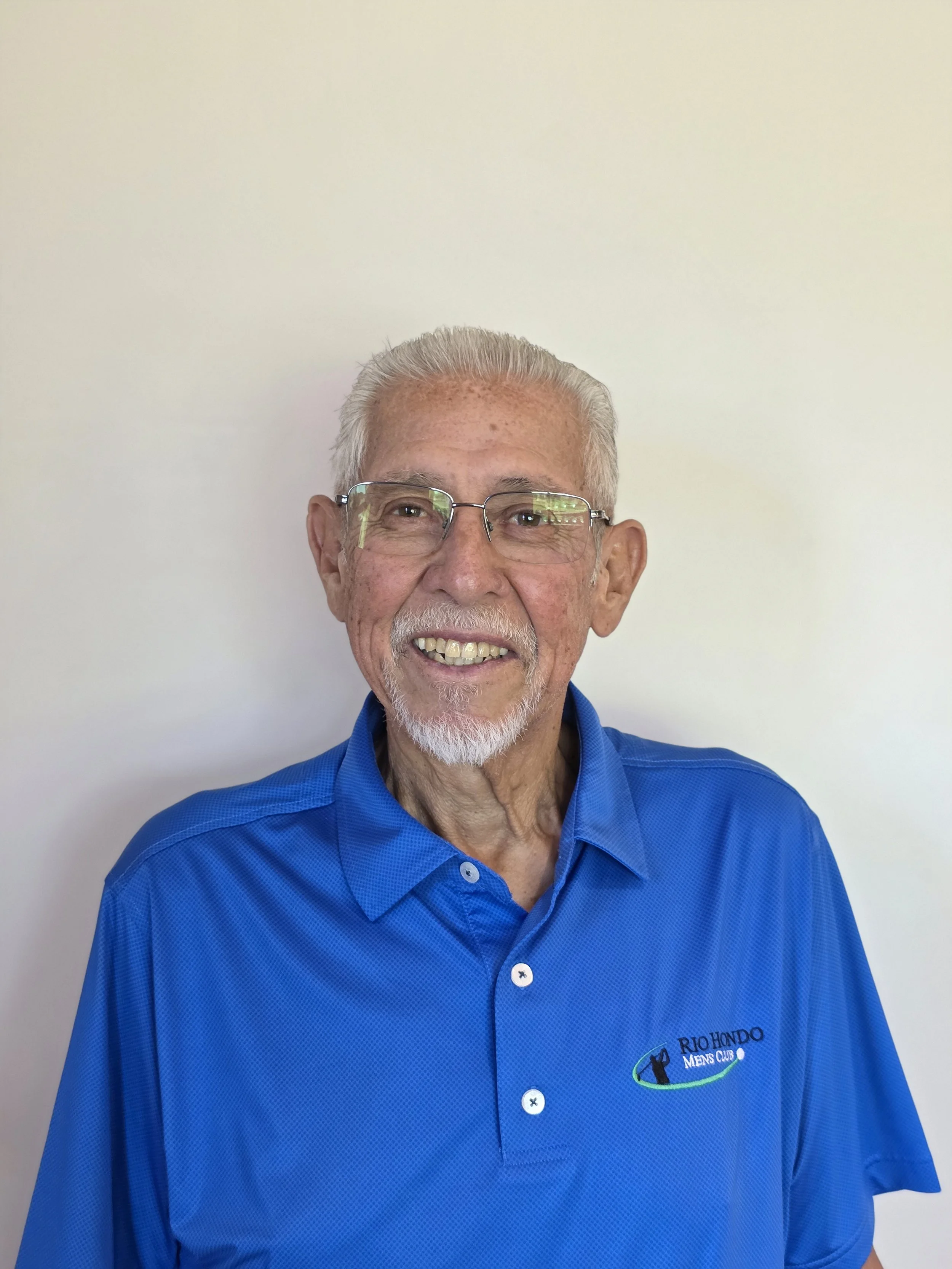 Elderly man with glasses, wearing a black "Rio Hondo Men's Club" polo shirt, smiling against a neutral background.