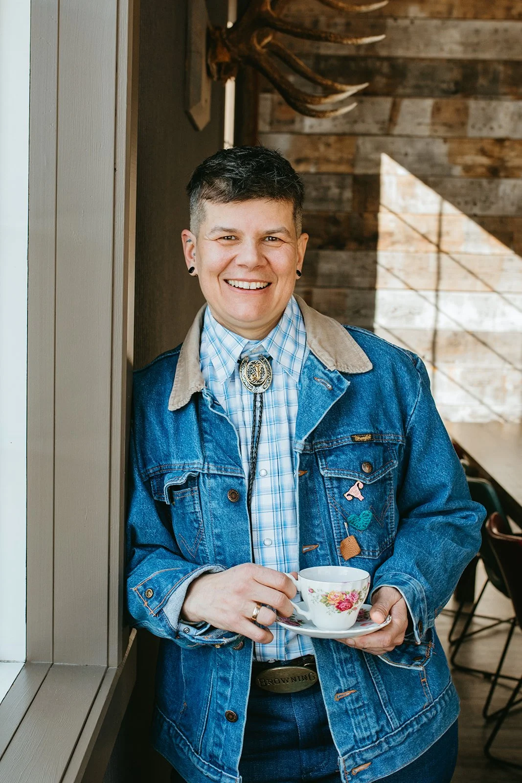 A smiling woman with short dark hair, wearing a denim jacket with patches and a bolo tie over a plaid shirt, holding a floral teacup and saucer, standing indoors near a window with wooden wall decor.