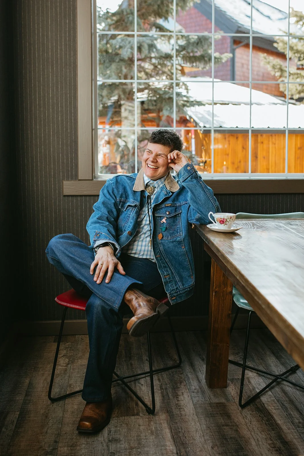 A man sitting on a red chair at a wooden table, smiling and laughing, wearing a denim jacket, plaid shirt, jeans, and cowboy boots, with a cup of tea on the table, inside a room with a large window showing trees and a red-roofed building outside.
