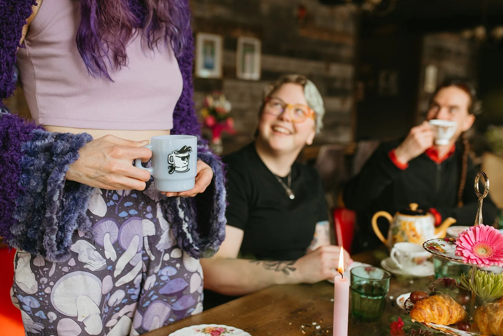 A woman holding a coffee mug with a cartoon face and sunglasses, wearing a purple fuzzy jacket and a patterned skirt, standing beside a table set for breakfast. Two people sit at the table, a woman with glasses holding a sparkler and a man drinking from a cup. The table has a teapot, flowers, and food items, and the background is a cozy wooden interior.