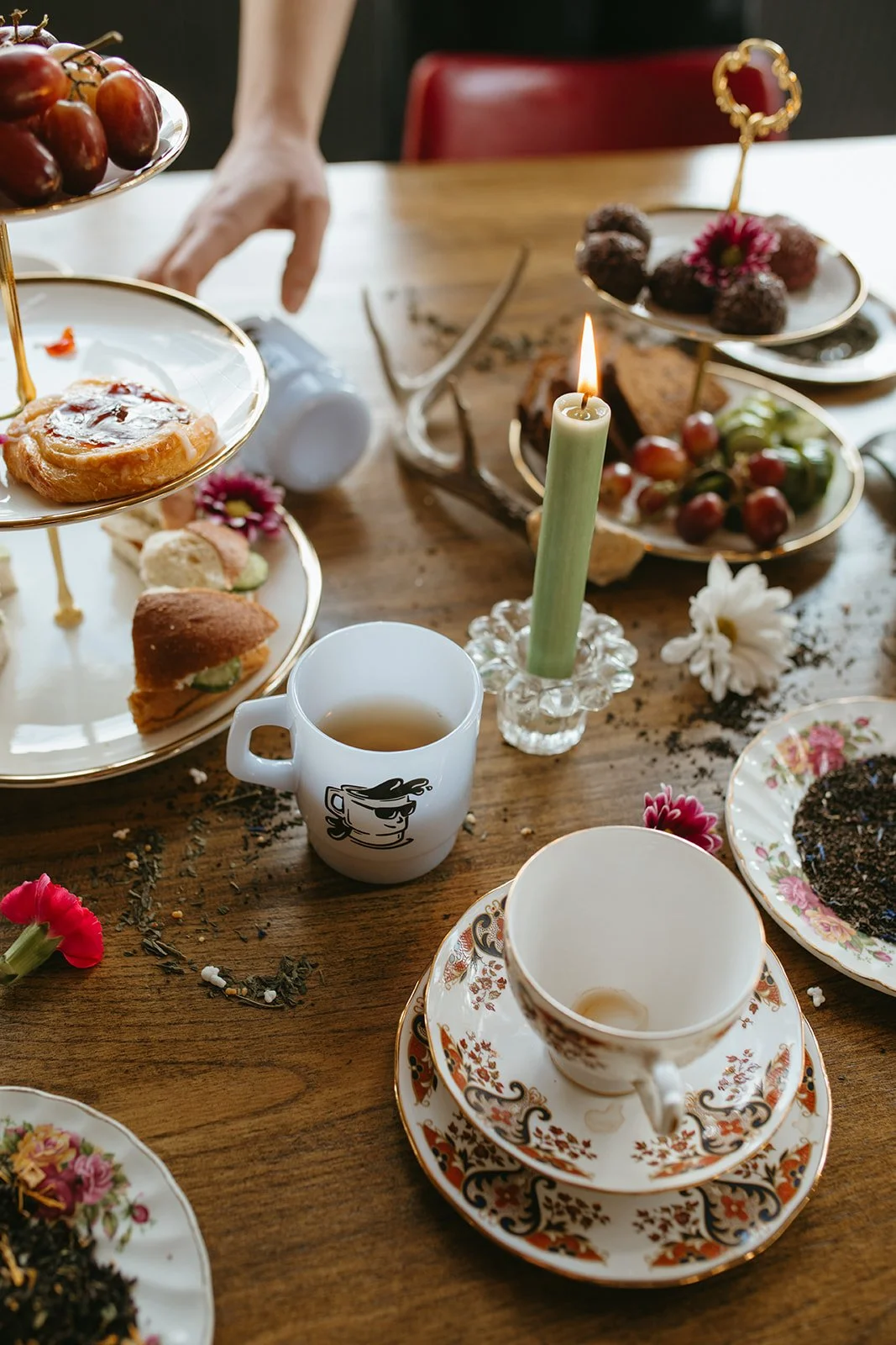 A wooden table set for tea with assorted snacks, teacup, candles, flowers, and a tiered dessert stand.
