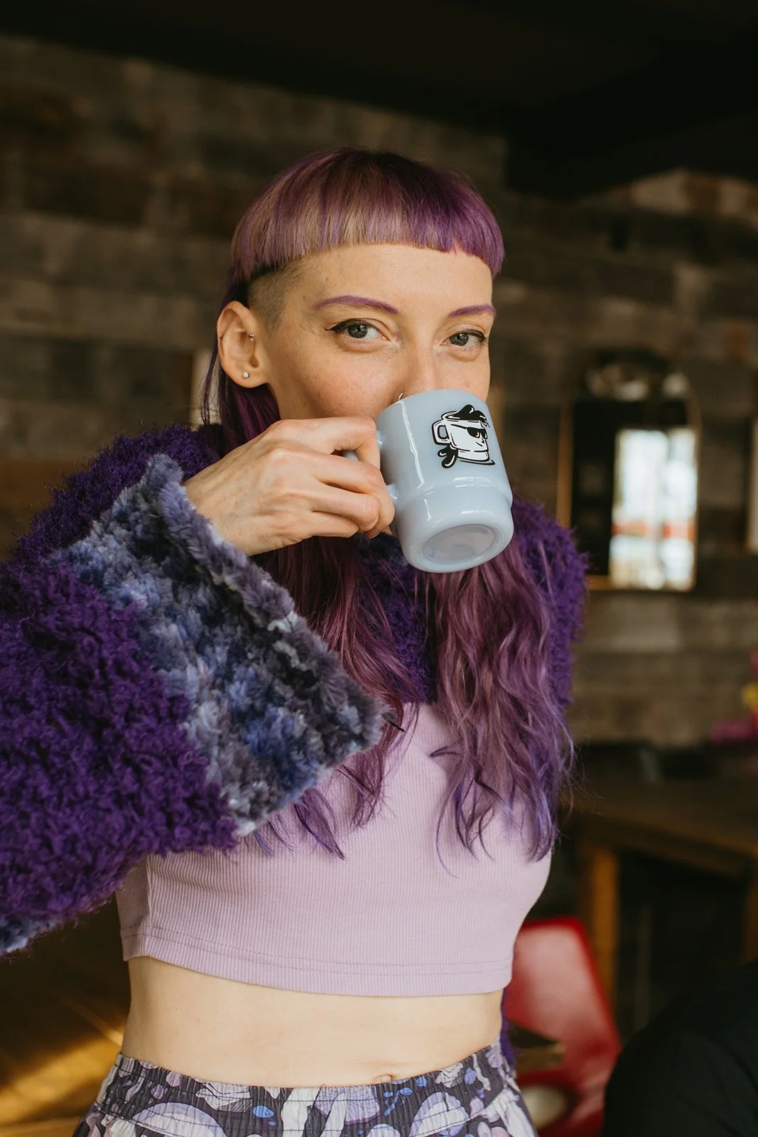 A woman with purple hair sipping from a mug with a cartoon coffee cup wearing sunglasses, inside a cozy wooden cafe.