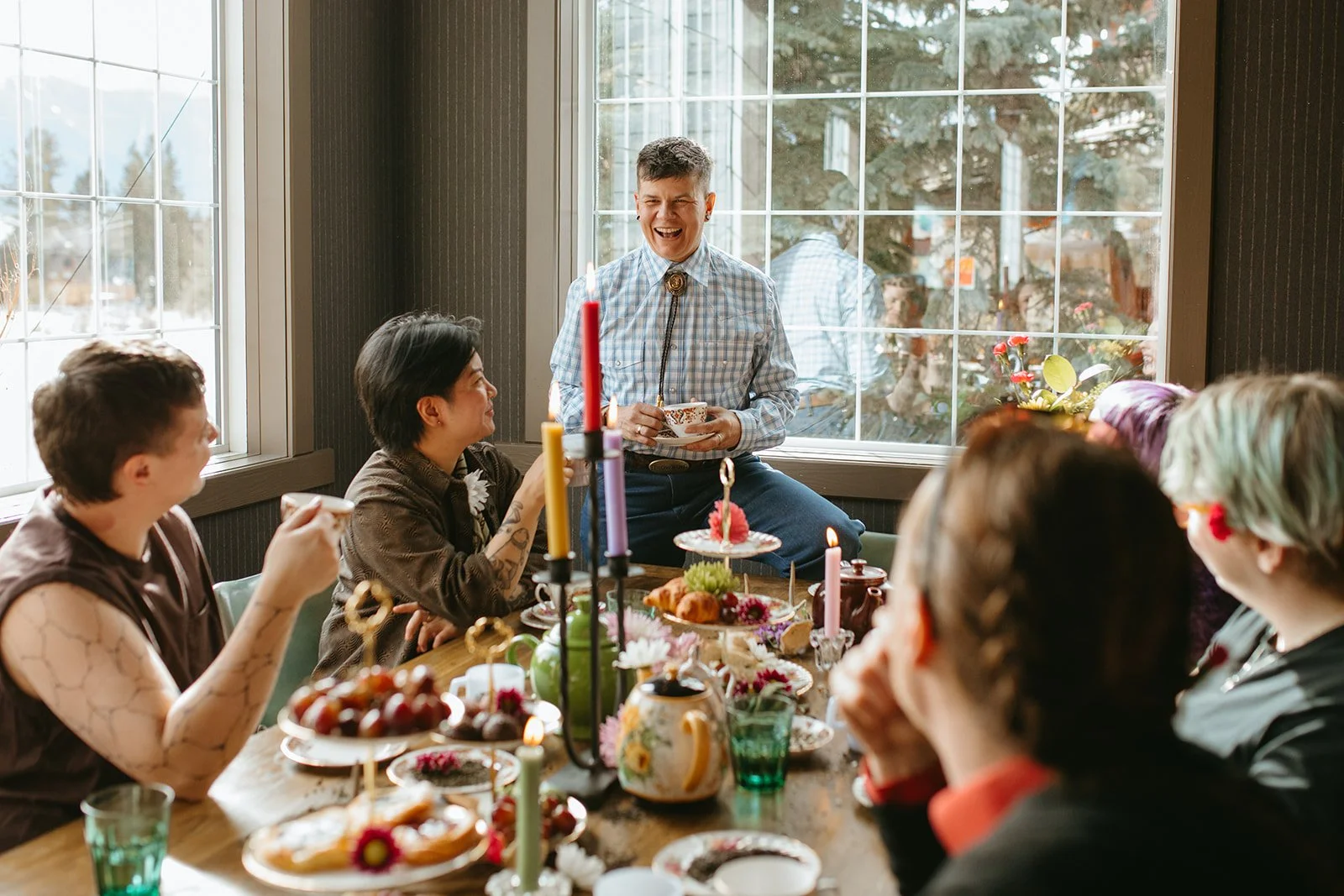 A group of people celebrating a birthday or special occasion at a dining table with food, drinks, and candles, with a man standing and smiling.