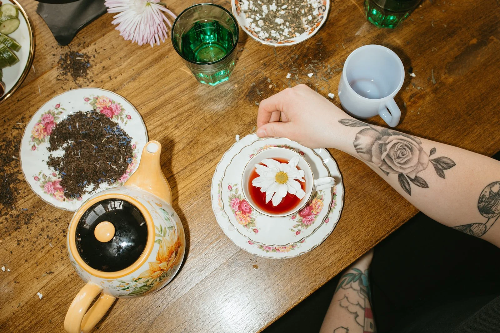 A wooden table set with a floral teapot, a floral teacup with a daisy floating in tea, a white mug, a bowl of mixed herbs or tea leaves, a plate of loose tea leaves, and two glasses of green-colored beverage. A person's arm with tattoos, including a rose and other designs, is reaching for the teacup.