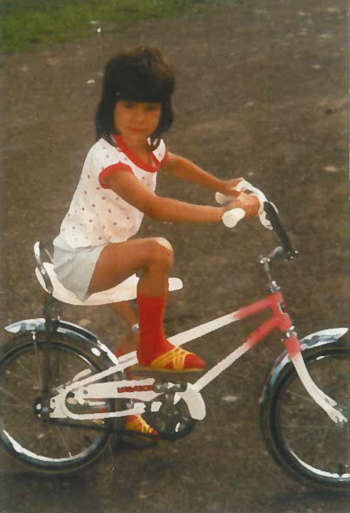 A young person with dark hair riding a pink and white bicycle outdoors on a dirt path, wearing a white dress with red trim, red socks, and yellow shoes.