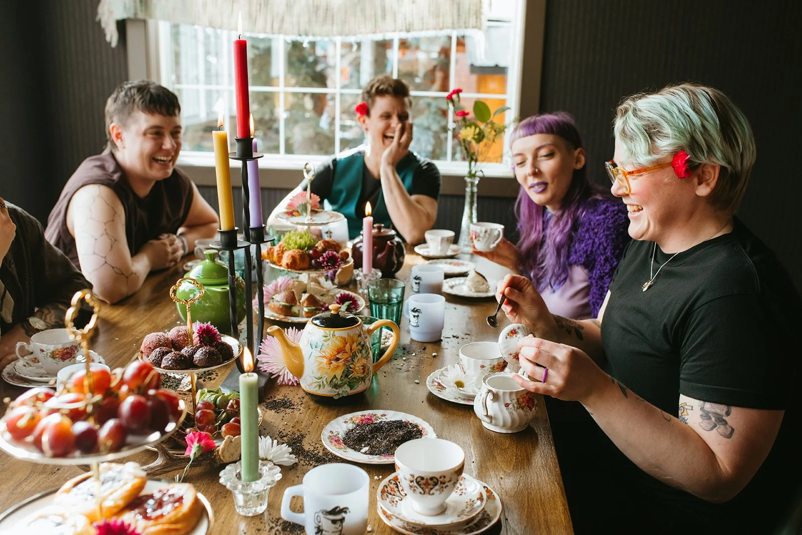 Group of friends enjoying a tea party with desserts, tea cups, and candles, sitting around a wooden table in a cozy room.