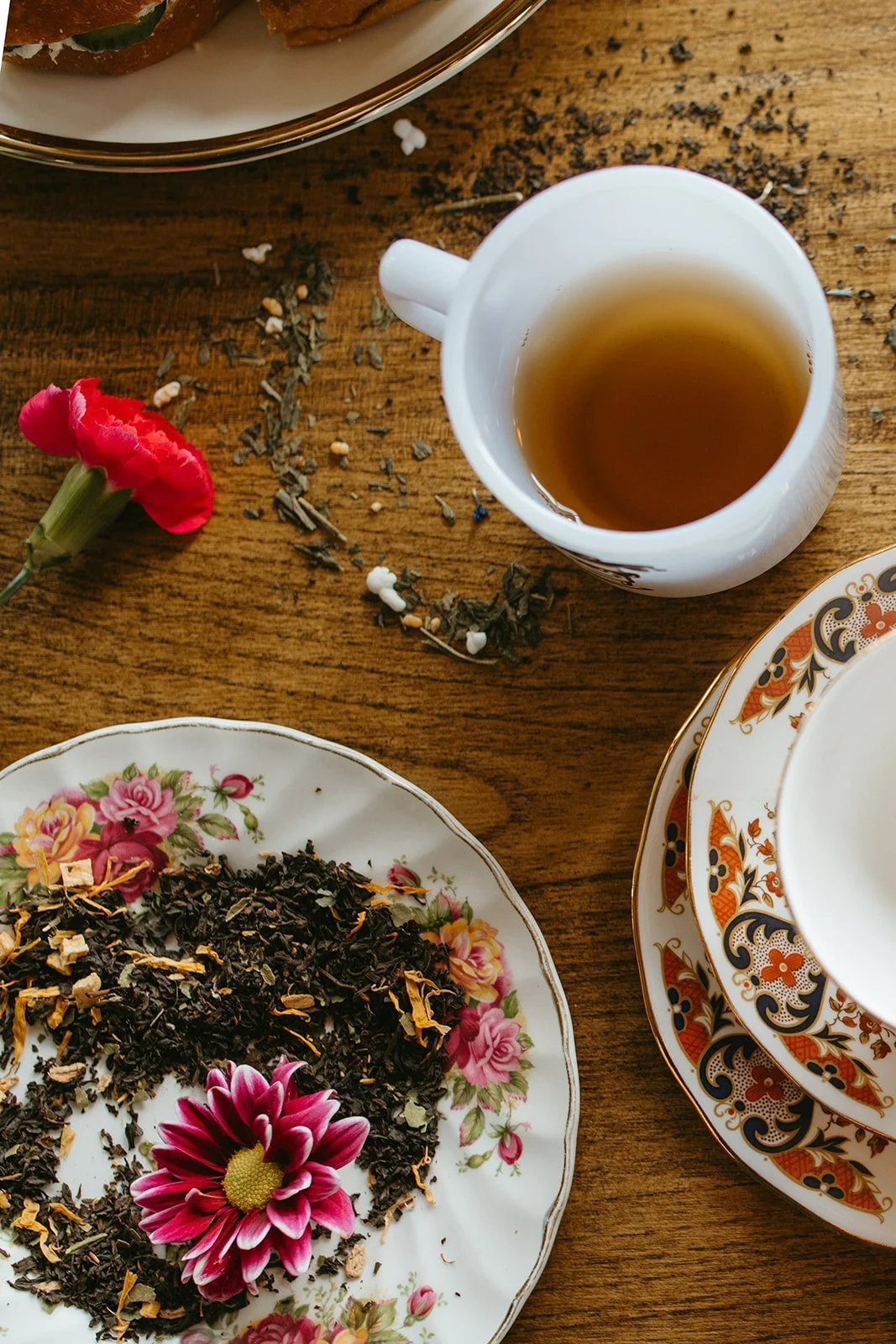 A table set with a floral plate of loose tea, a white mug of brewed tea, a decorative cup and saucer, a second plate with a pink flower, and a floral dish with more loose tea. There is a small pink flower on the table and some tea leaves scattered around.