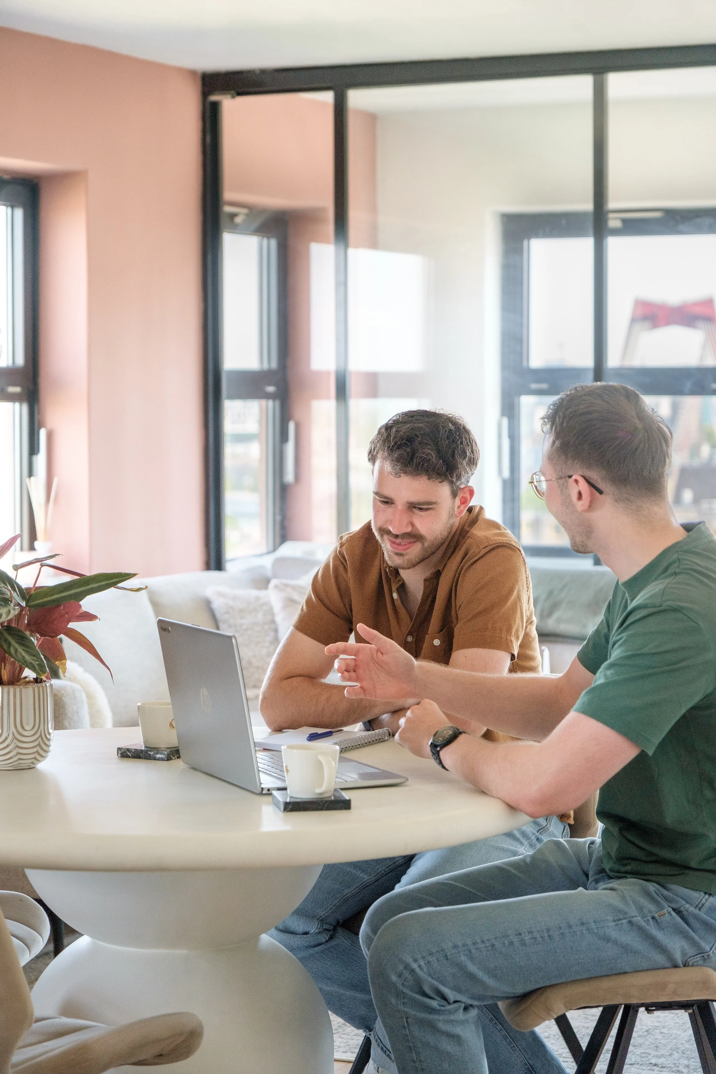 De trainer en klant die samen aan een tafel in een woonkamer werken met een laptop en een bak koffie.