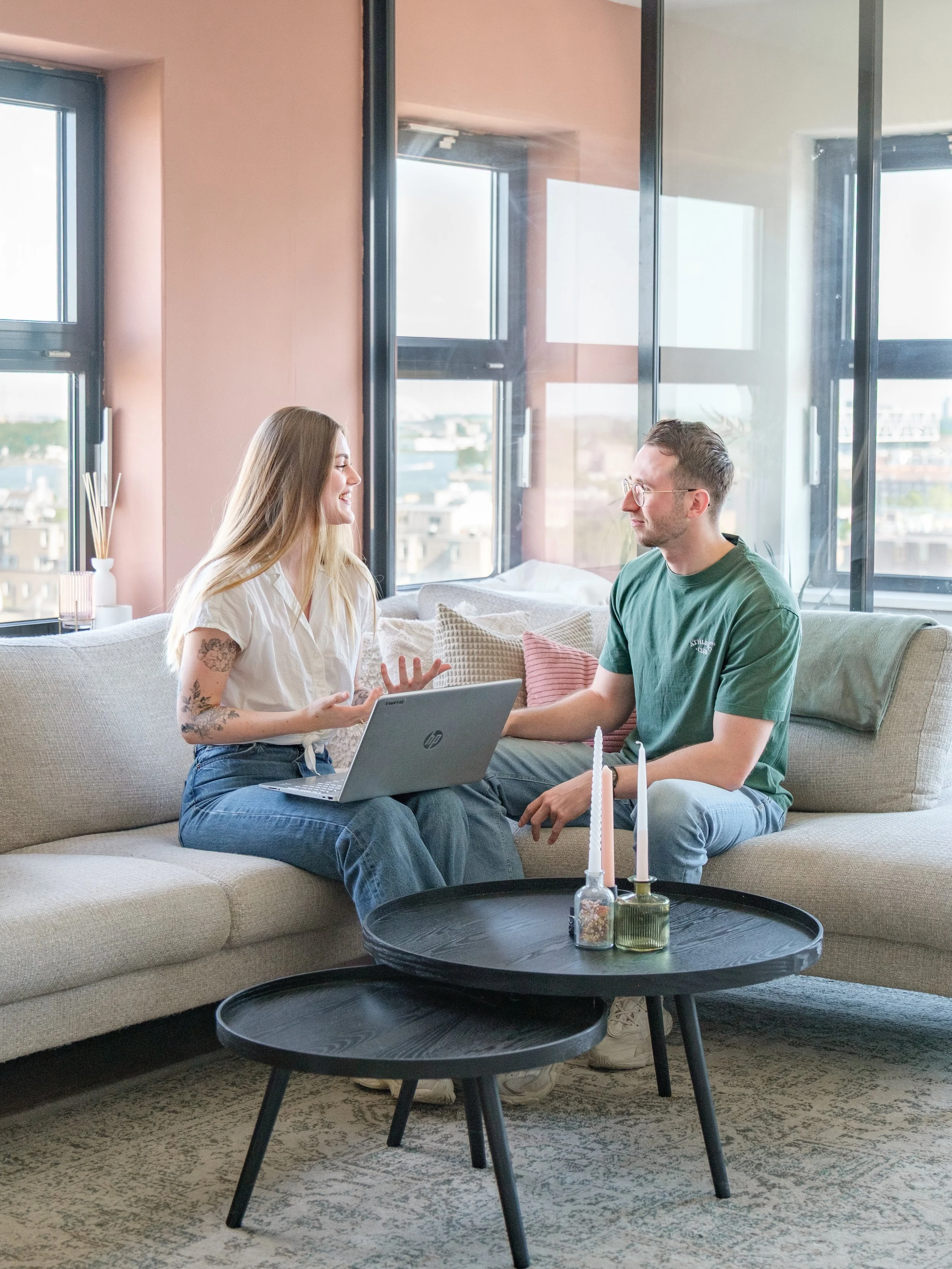 De trainers zitten op een bank in een lichte woonkamer met grote ramen en praten, terwijl de vrouw een laptop vasthoudt.