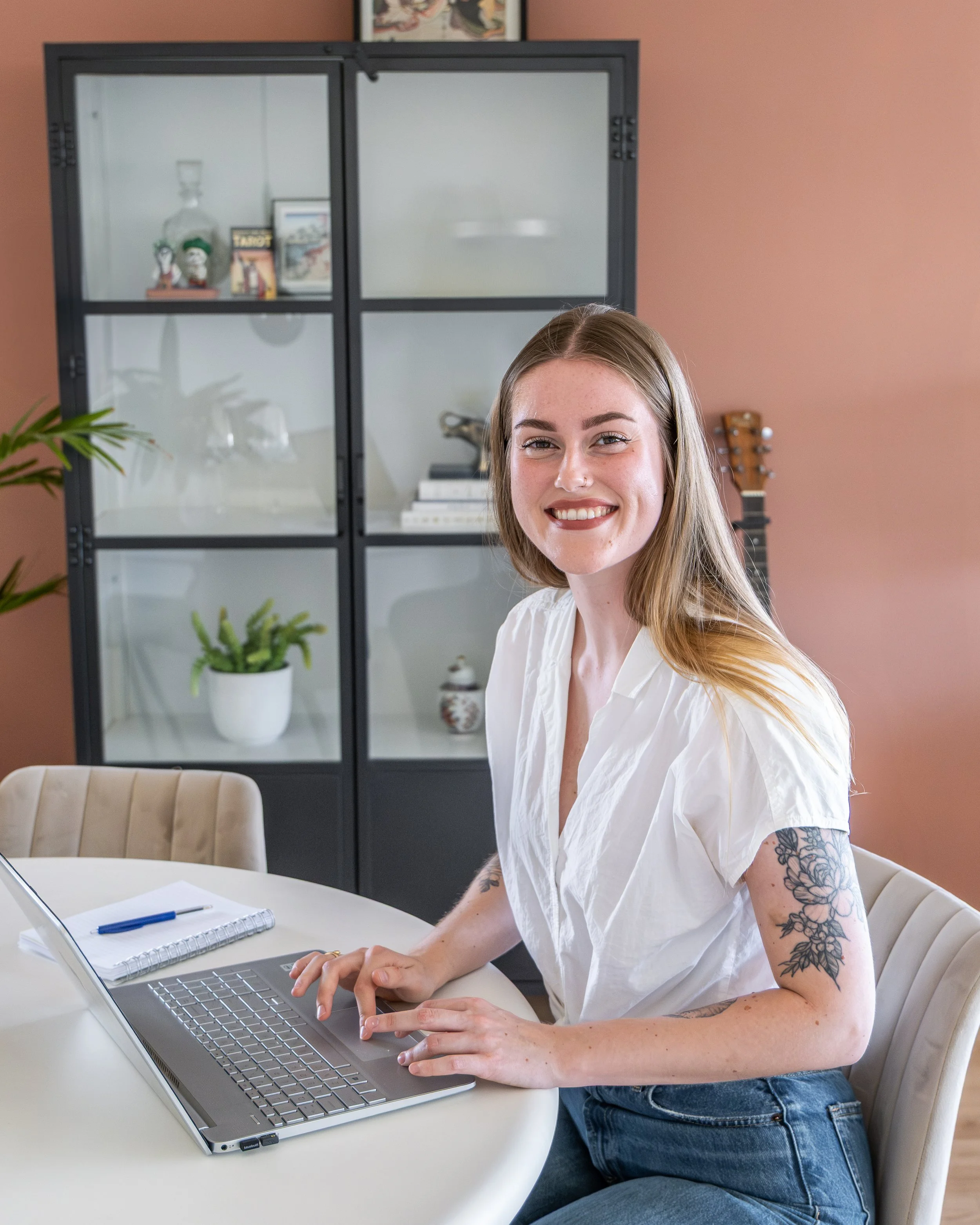 Een lachende vrouw met lang, lichtbruin haar zit aan een witte tafel, werkt op een laptop, in een moderne kamer met een glazen kast en een vissenplant.
