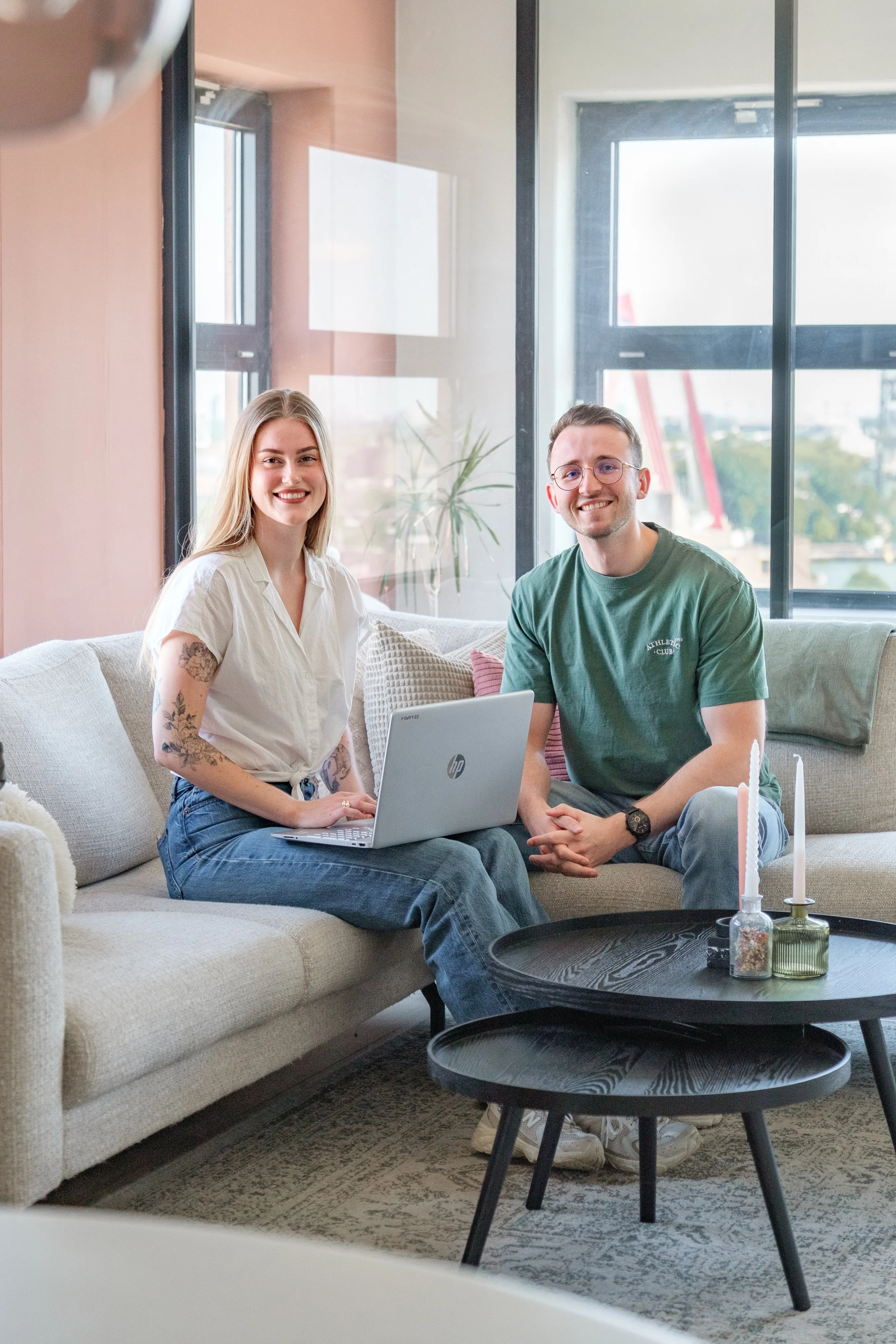 Twee jonge mensen zitten op een bank, werken met een laptop, in een modern, licht interieur met grote ramen en planten.