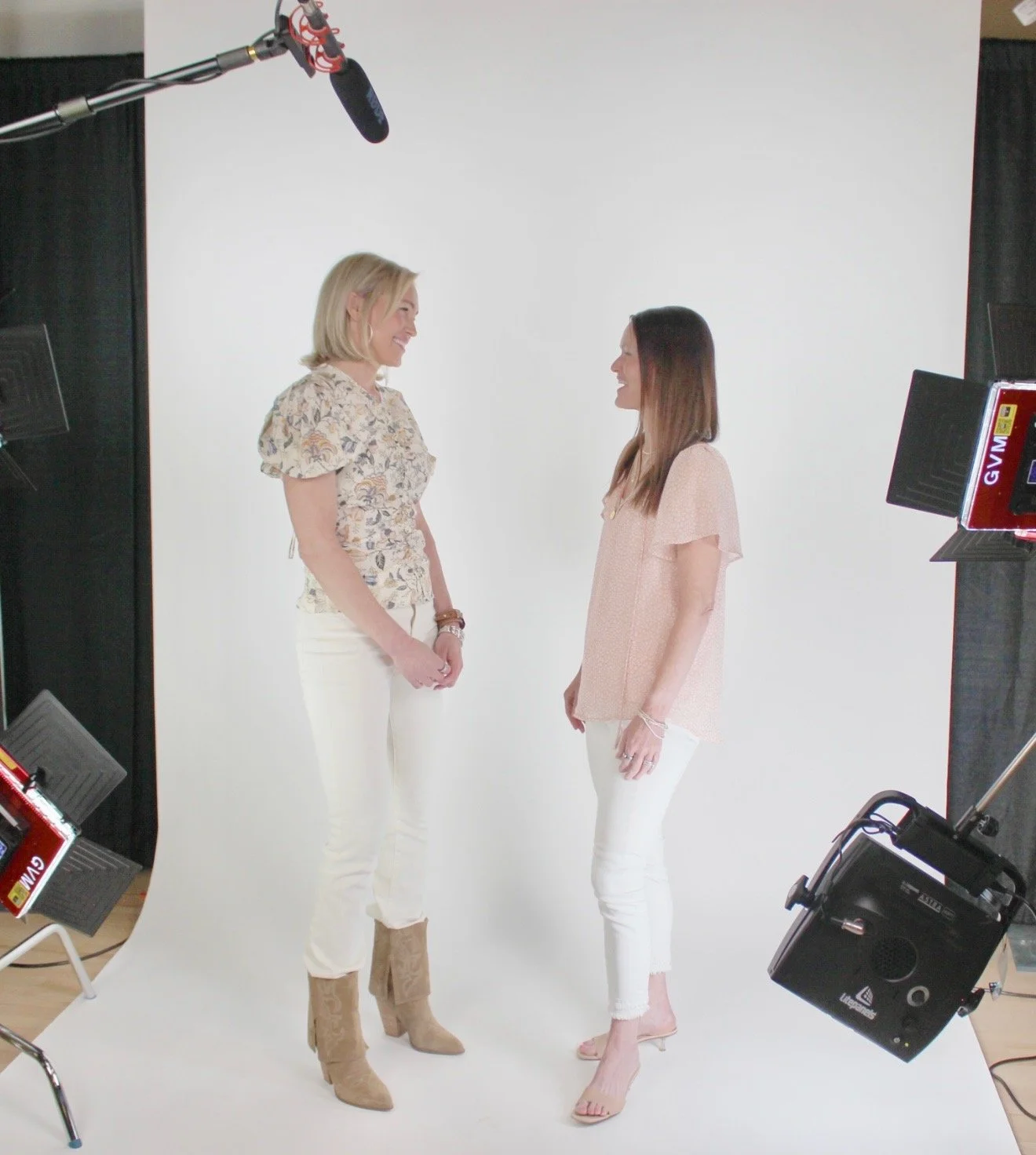 Two women in light-colored clothing smiling and opposite each other in a photography studio with a white backdrop and professional lighting equipment.
