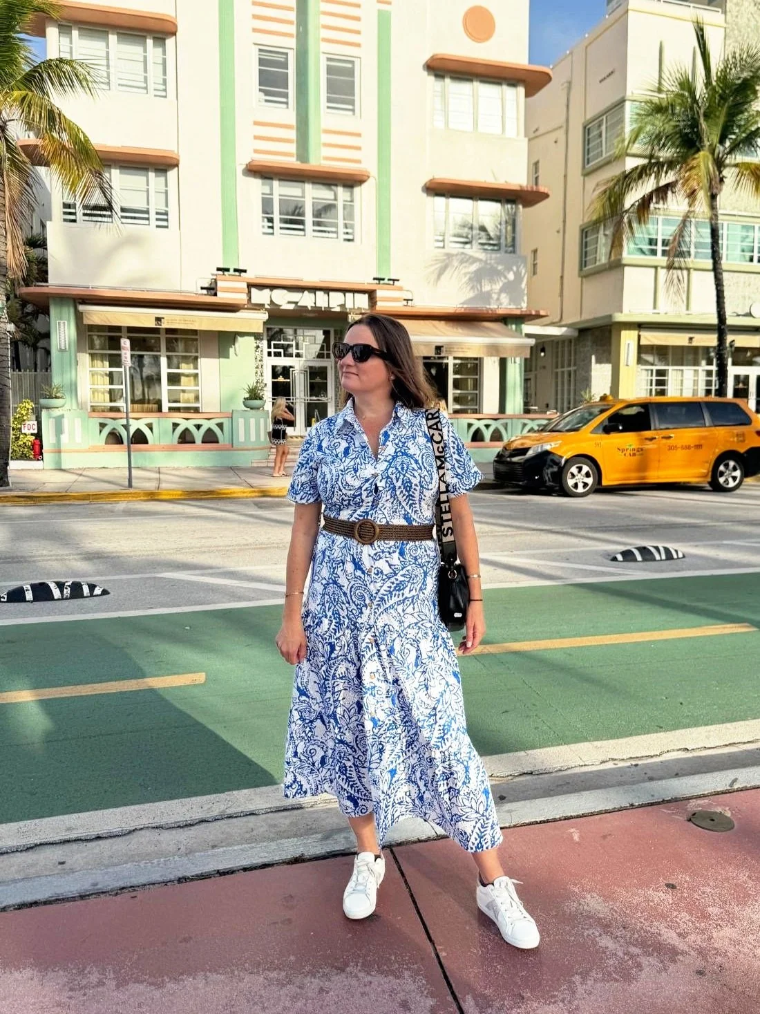 A woman in a white and blue patterned dress, wearing sunglasses and white sneakers, standing on a colorful crosswalk in front of pastel-colored buildings with palm trees. She has a black bag and a brown belt.