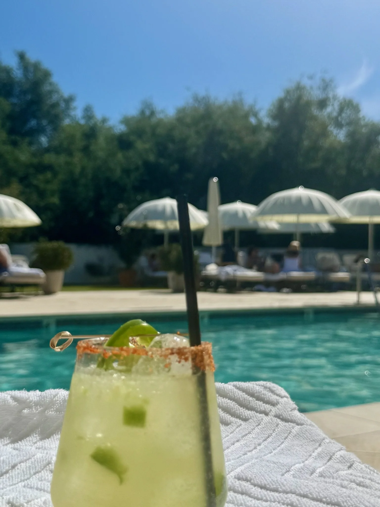 Poolside drink with lime and spicy rim on a white towel near a swimming pool, surrounded by lounge chairs and umbrellas.