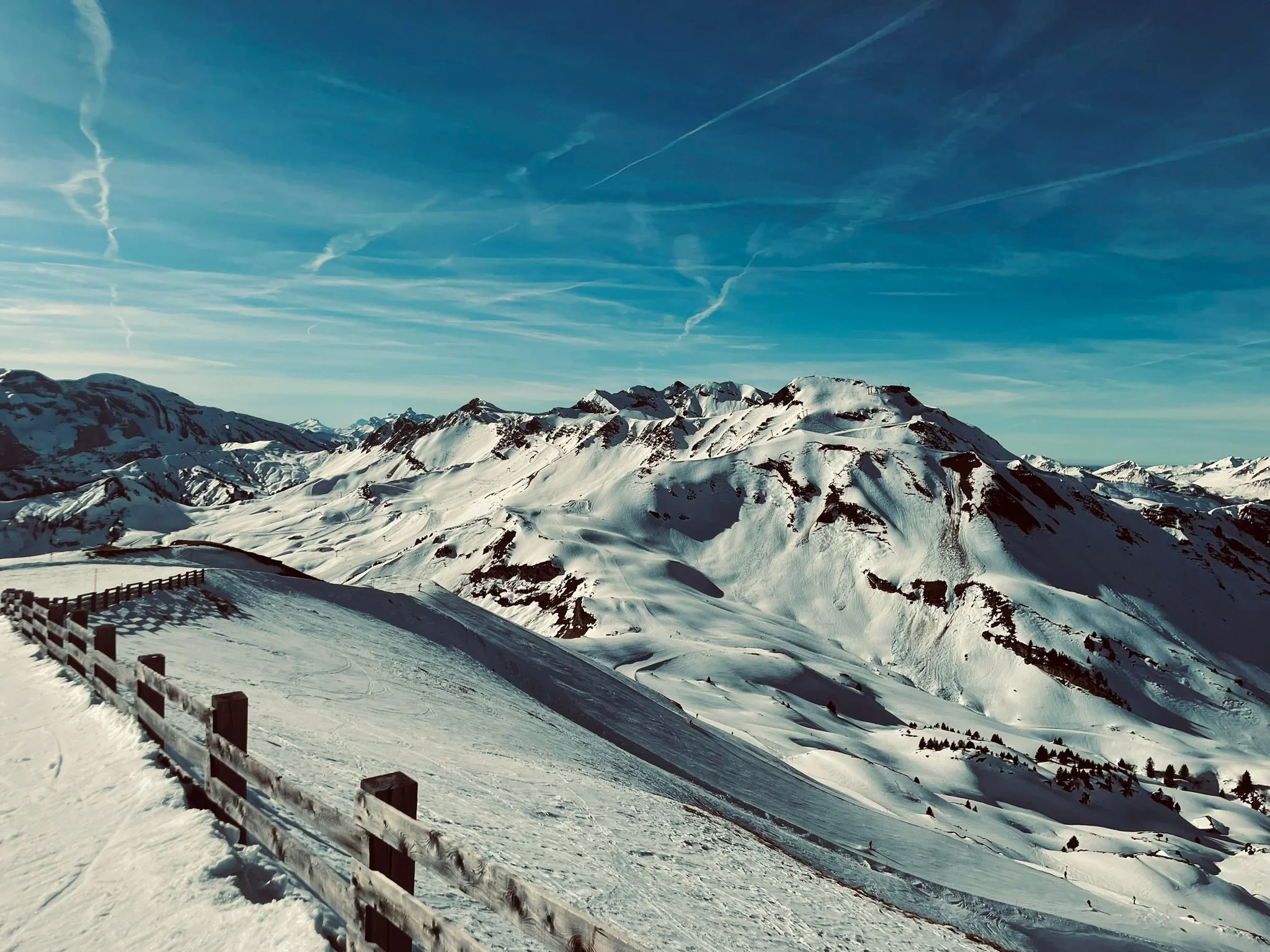 Snow-covered mountain landscape with a wooden fence along a slope. Clear blue sky with visible contrails.