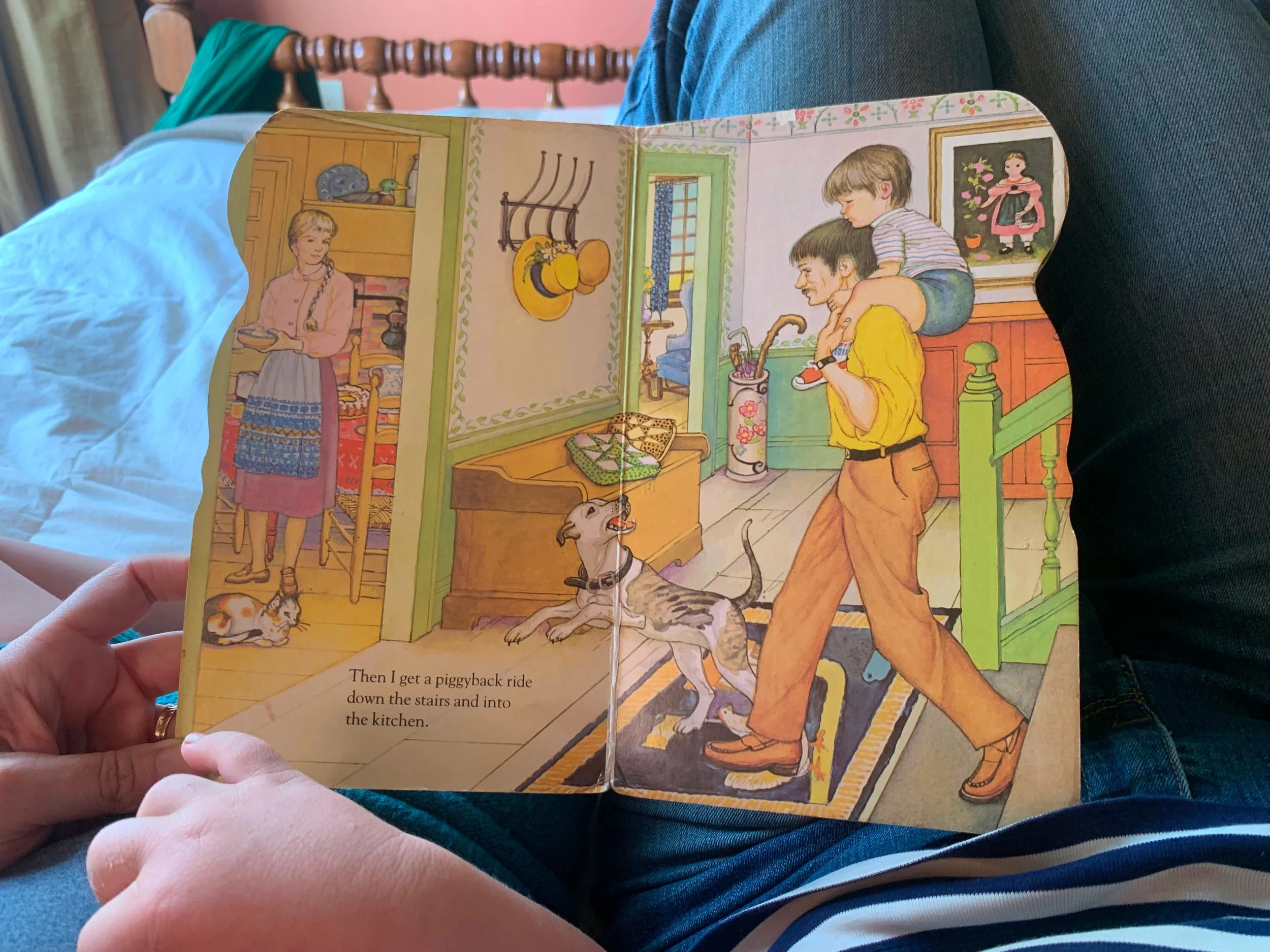 A colorful illustrated children's book depicts a boy giving a piggyback ride into the kitchen. Inside, a dog is sitting on the floor. A woman with a braid stands in a doorway holding a plate. The kitchen has a patterned rug, hooks with hats on the wall, and a window with curtains.