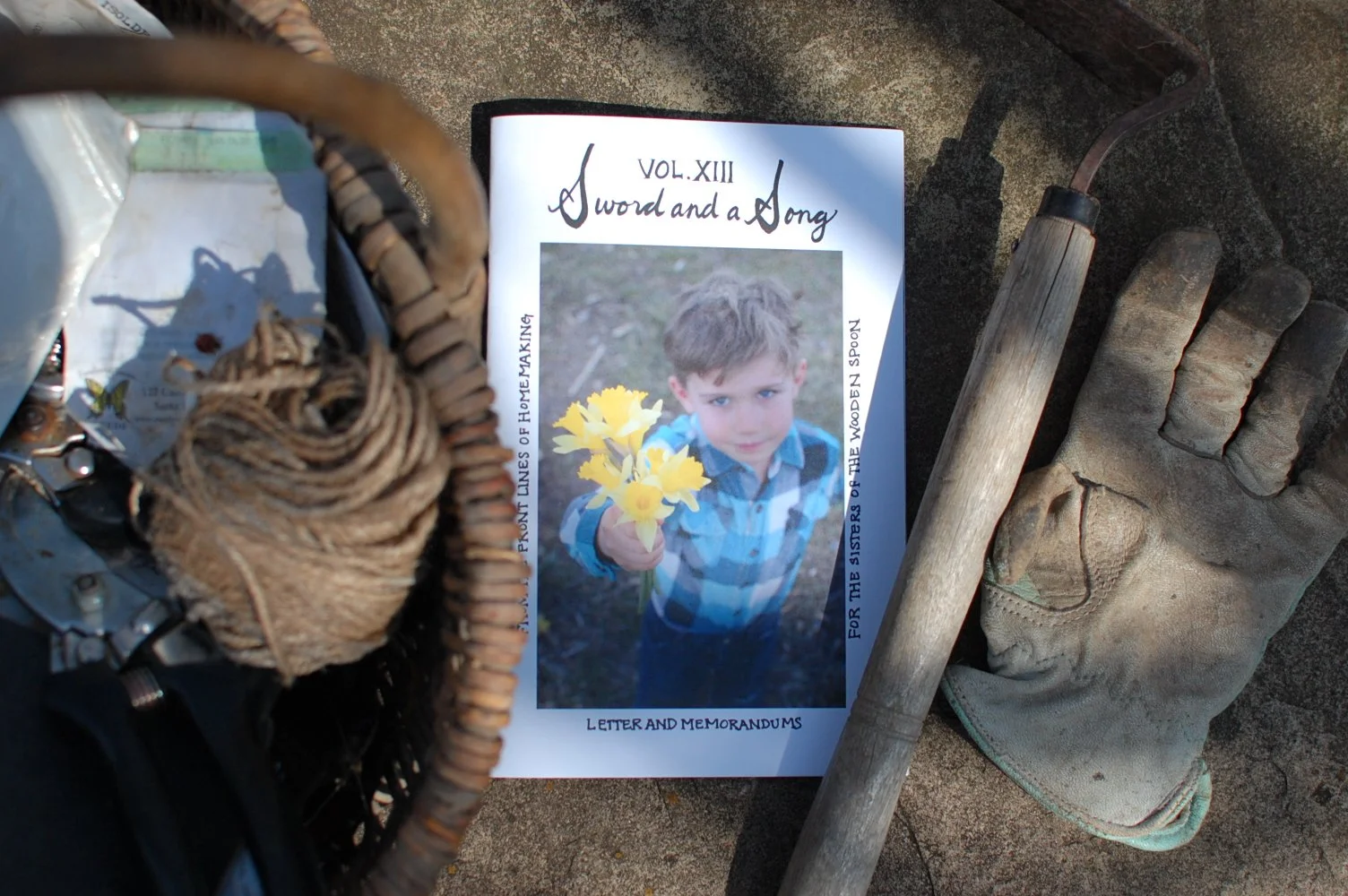 A magazine titled 'A Sword and a Song' with a photo of a young boy holding yellow flowers, placed on a sandy surface next to a gardening glove, a wooden stick, and part of a basket and a shovel.