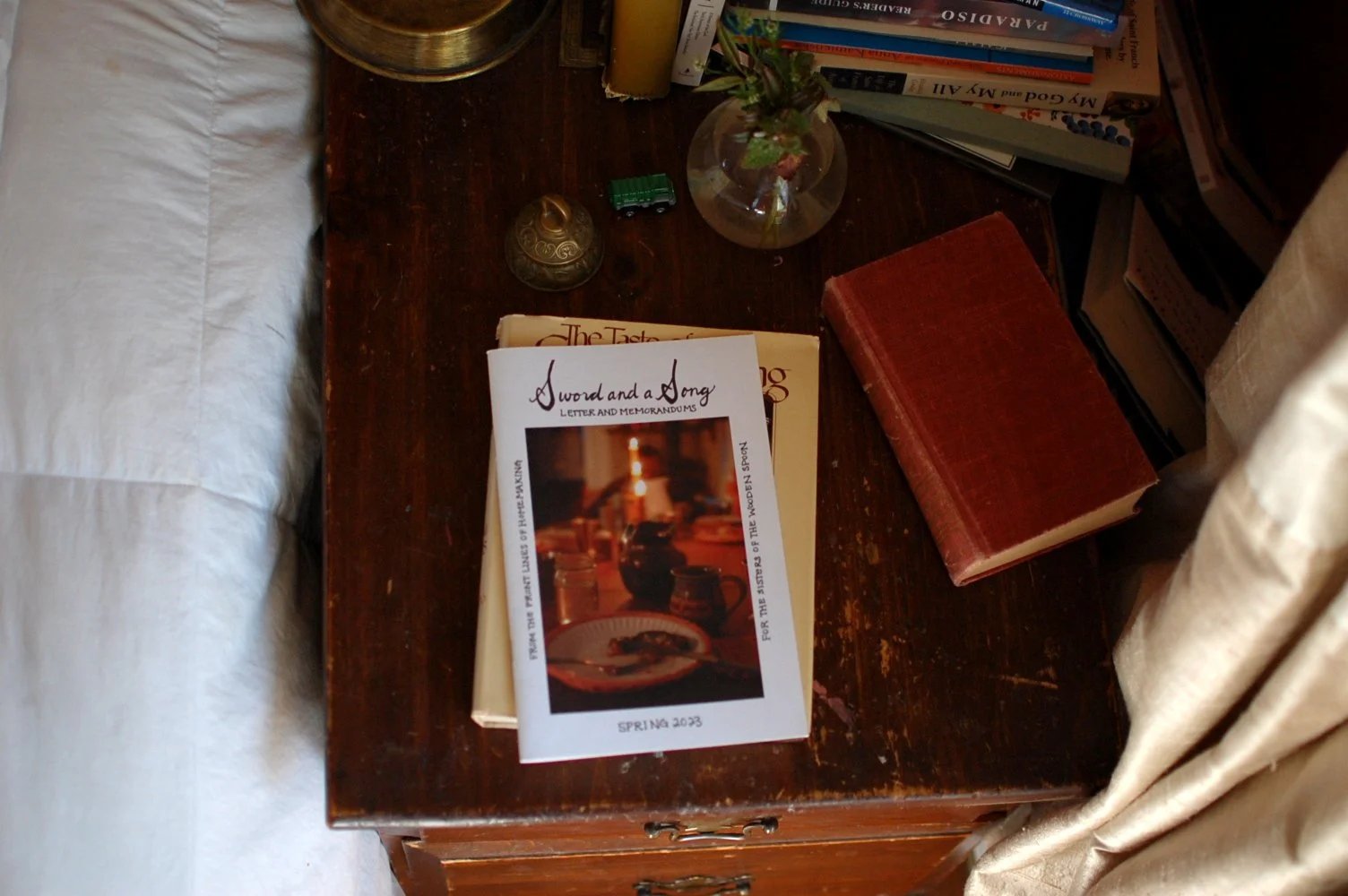 A wooden bedside table with books, a glass vase with flowers, a decorative metal item, and a printed program titled "A Sword and a Song" resting on top of stacked books.