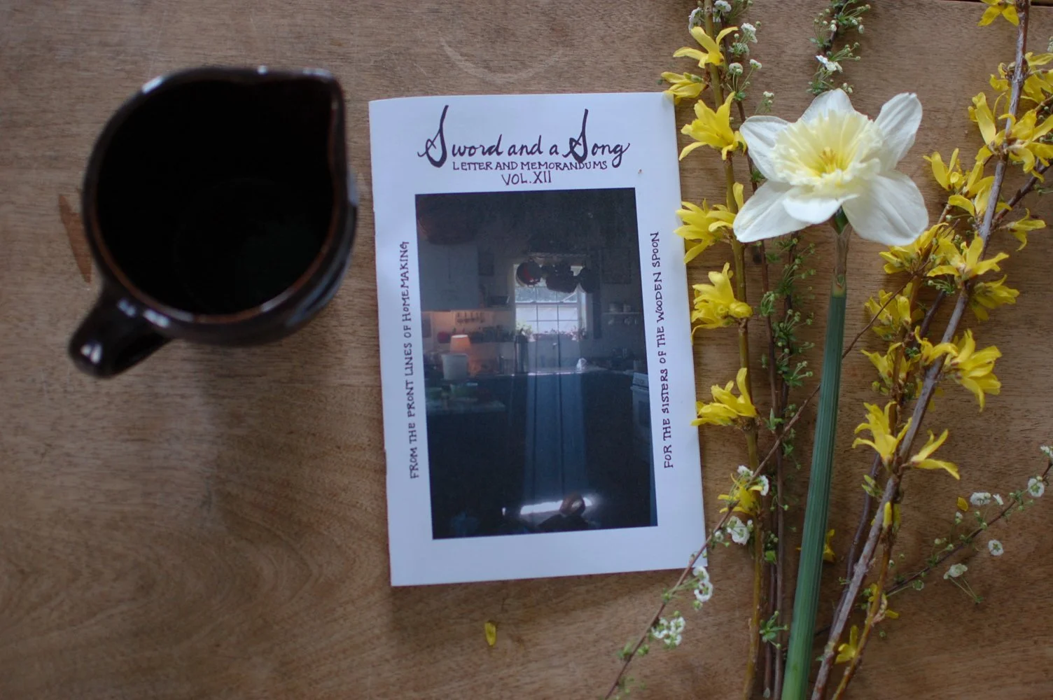 A wooden table with a dark coffee mug, a printed magazine titled "A Sword and a Song" with a photo of a windowed room, and a bouquet of yellow and white flowers.