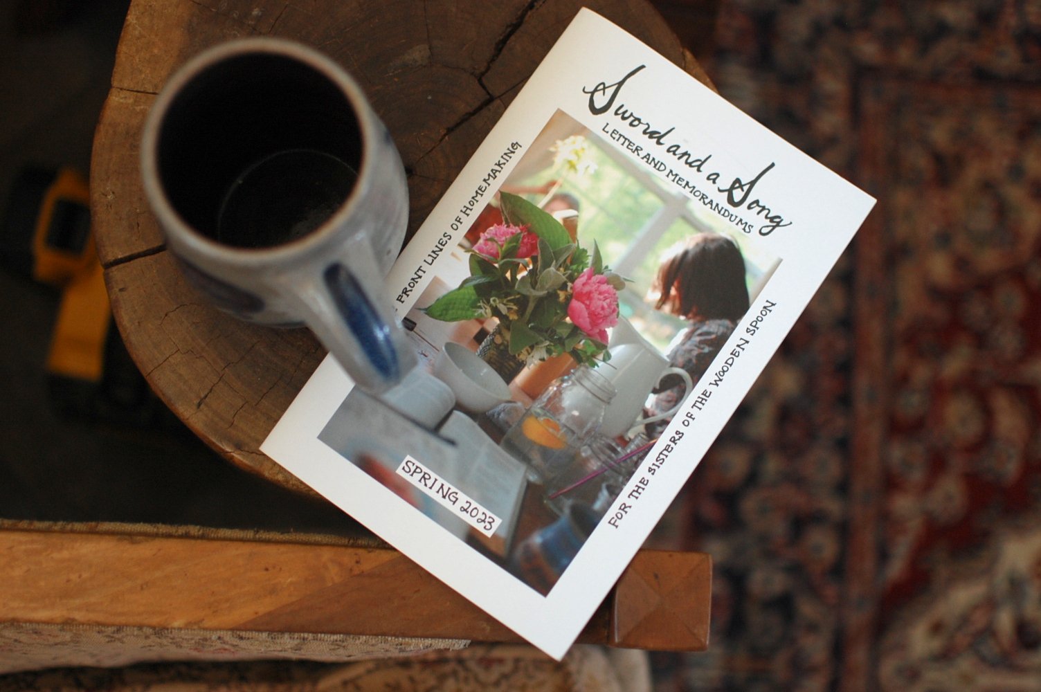 Coffee mug on a wooden table with a magazine featuring a flower and a woman in a bright room.