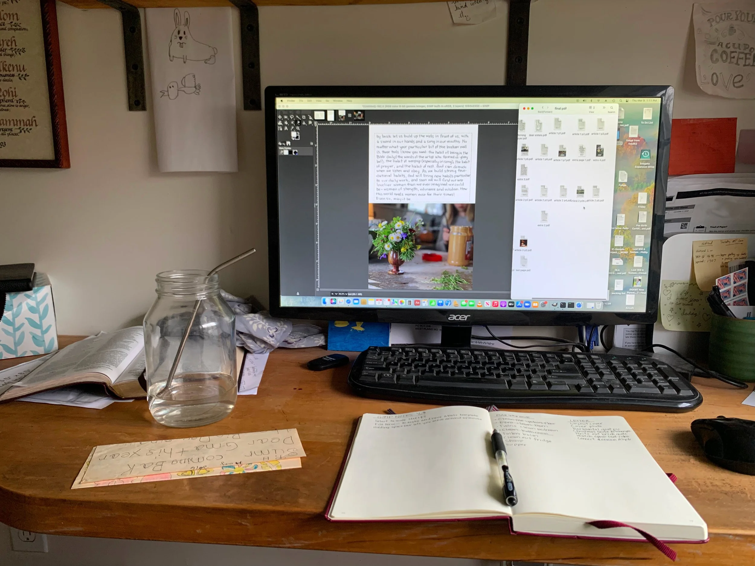 A cluttered wooden desk with an open book, a glass jar with a metal straw, a computer monitor displaying photo editing software, a black keyboard, a computer mouse, a pen in a notebook, and various papers and notes. The background shows a wall with drawings, notes, and motivational quotes.