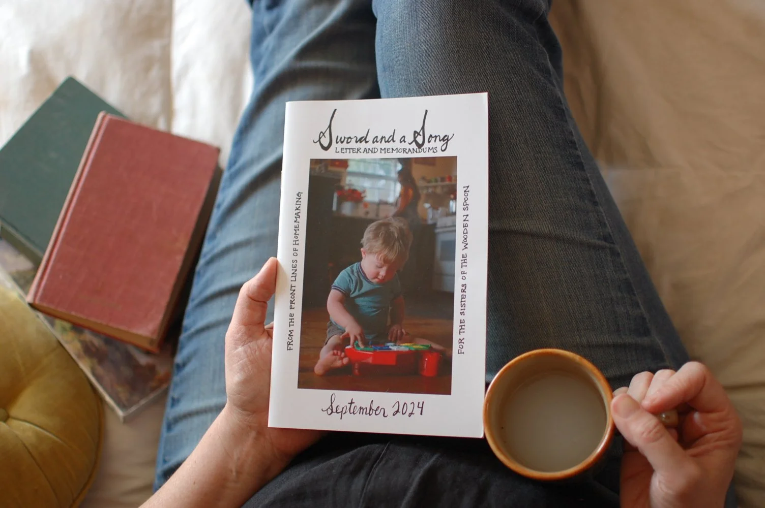 Person sitting on a couch holding a booklet with a photo of a young boy playing with a toy, a cup of coffee, and two books on a table beside them.