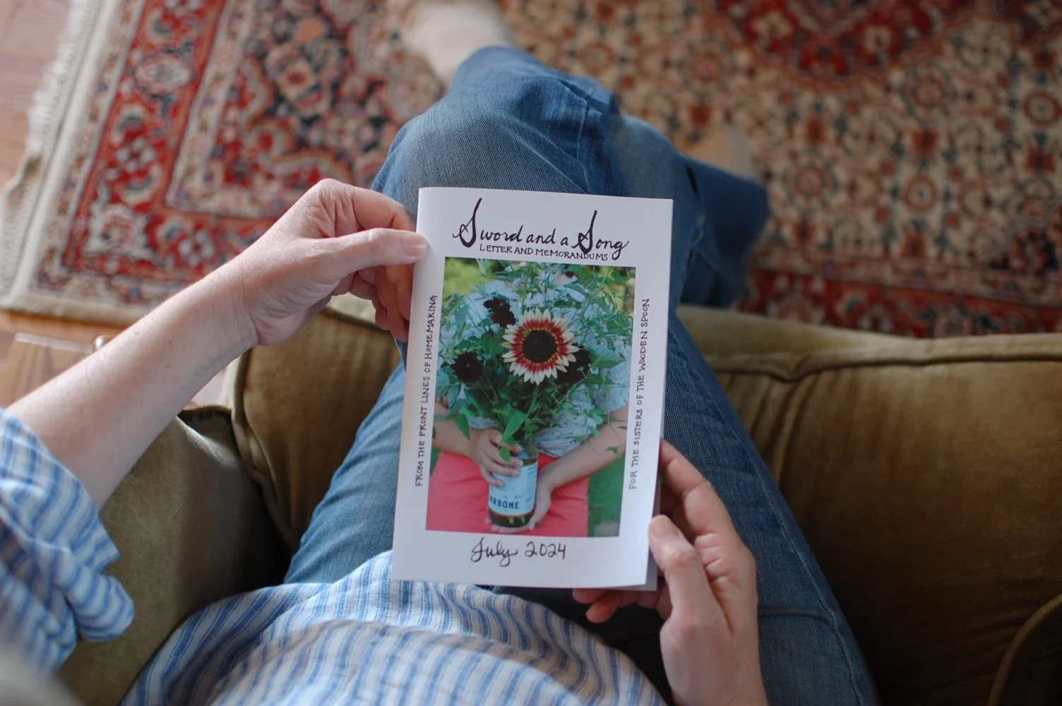 Person sitting on a couch holding a colorful brochure featuring a bouquet of flowers and a person. The brochure is titled "A Sword and a Song" and has the date July 2024 written at the bottom.
