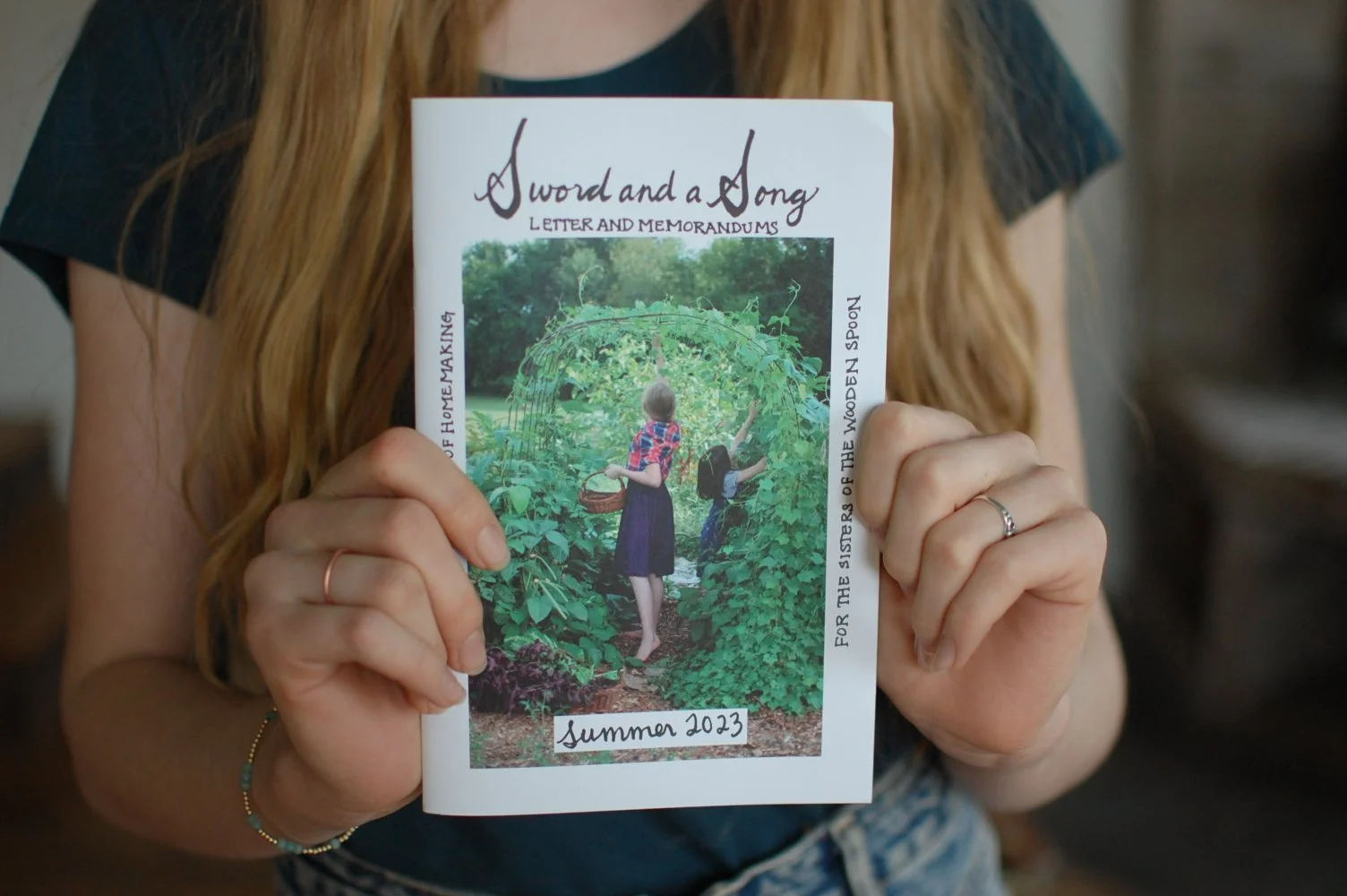 A person with long red hair holding a booklet titled 'A Word and a Song,' featuring a photo of two children picking berries in a garden on its cover. The booklet is labeled 'Summer 2023'.