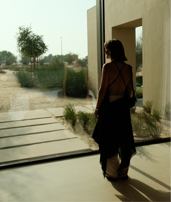 A woman with shoulder-length dark hair stands inside near a large glass window, looking outside at a desert landscape with sparse trees and bushes.