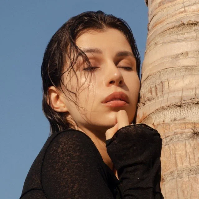 Close-up of a woman with wet hair and closed eyes, touching her chin with her hand, outdoors next to a palm tree against a blue sky.