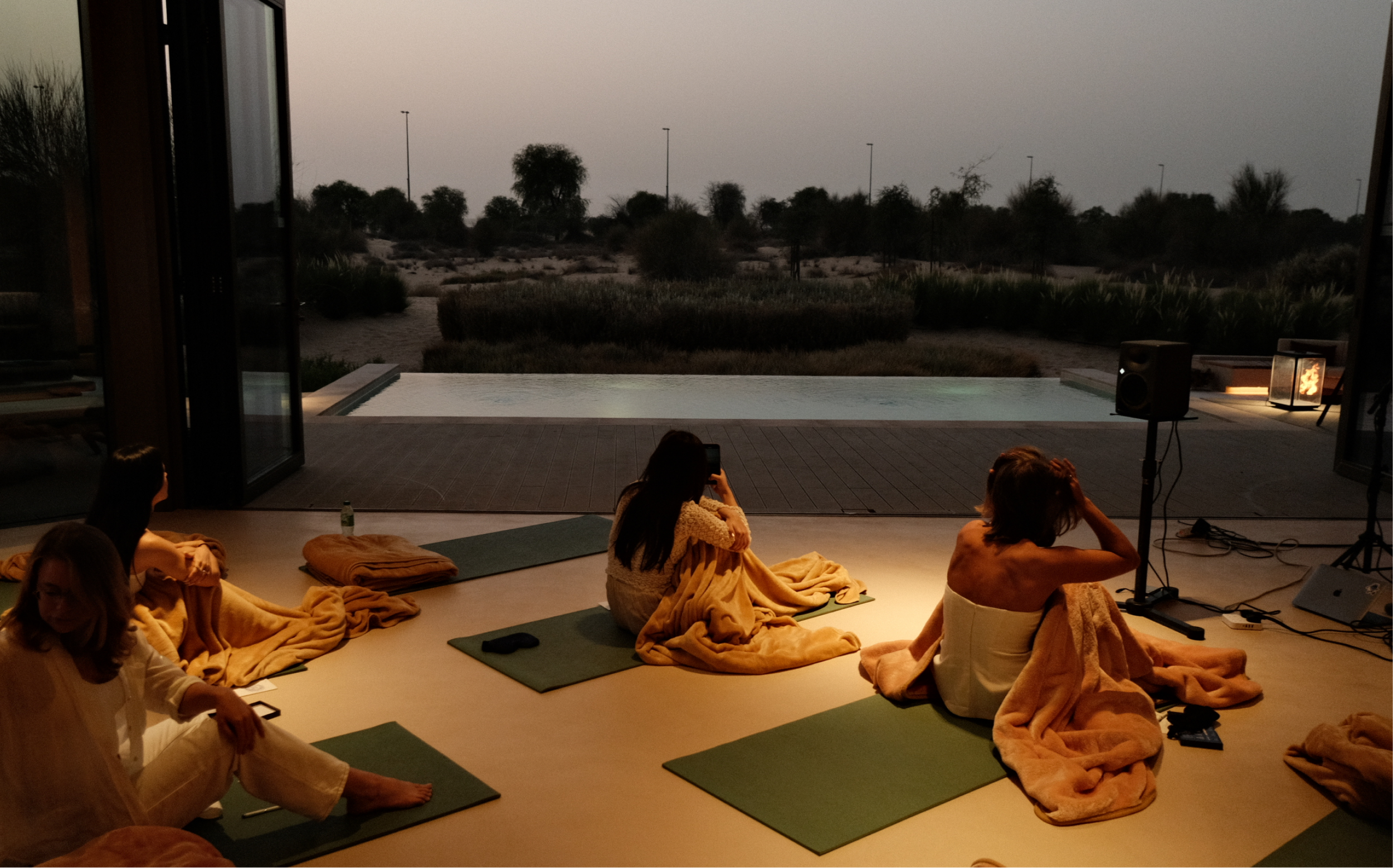 People seated on yoga mats and blankets in a dimly lit room with large open doors revealing an outdoor desert landscape with sparse vegetation and a pool, during sunset.