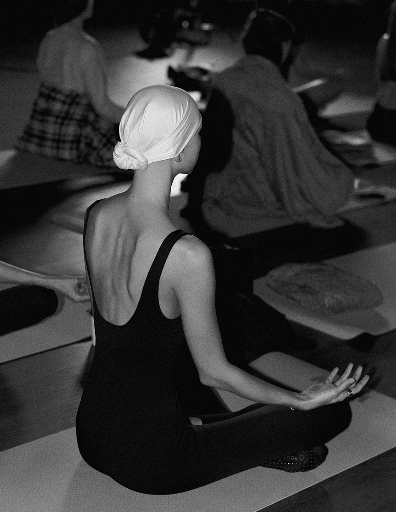 A woman practicing yoga in a seated meditation pose, wearing a headscarf and a sleeveless top, in a group setting with others in the background.